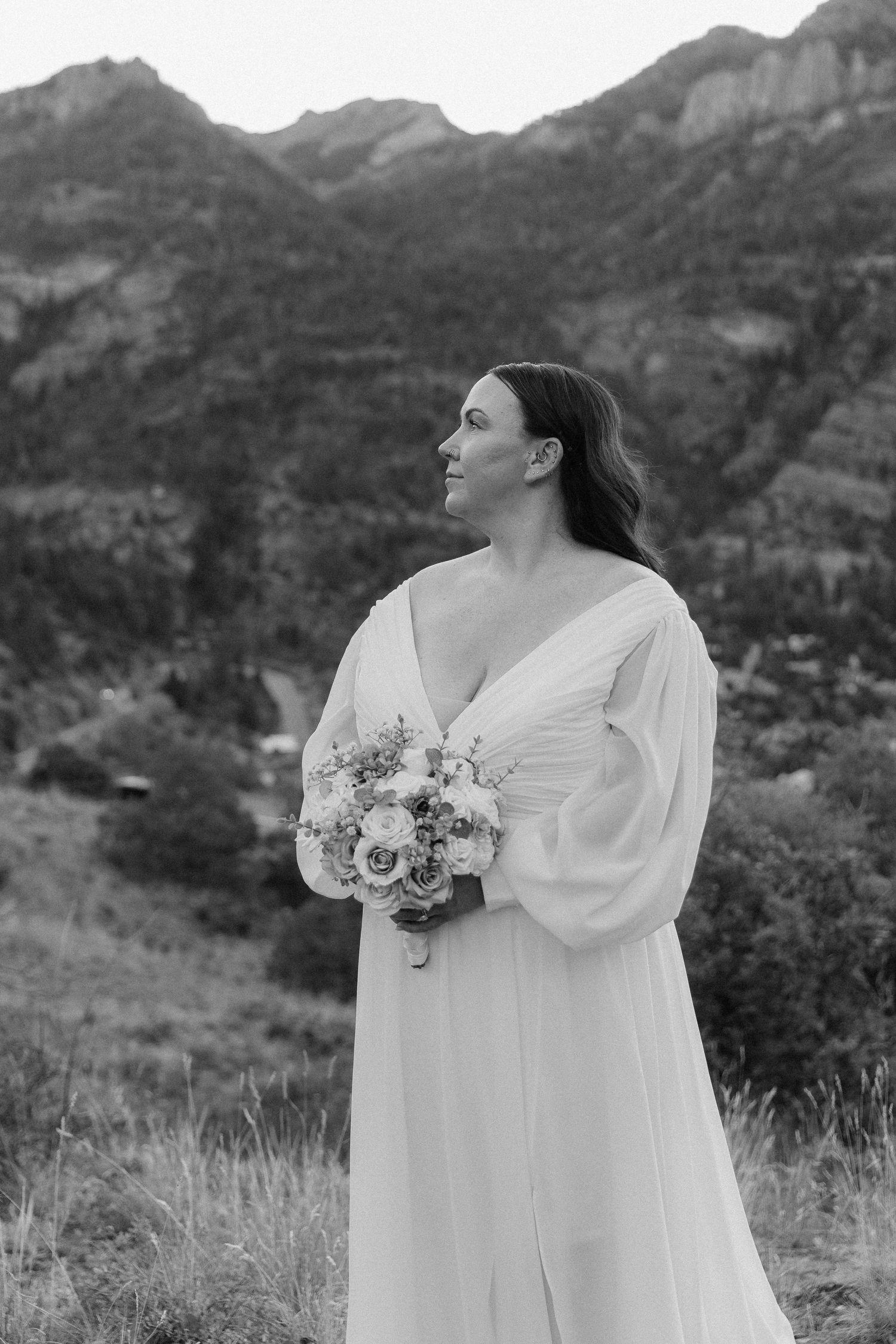 Bride in white gown holding floral bouquet poses before mountain backdrop in elegant black and white portrait.