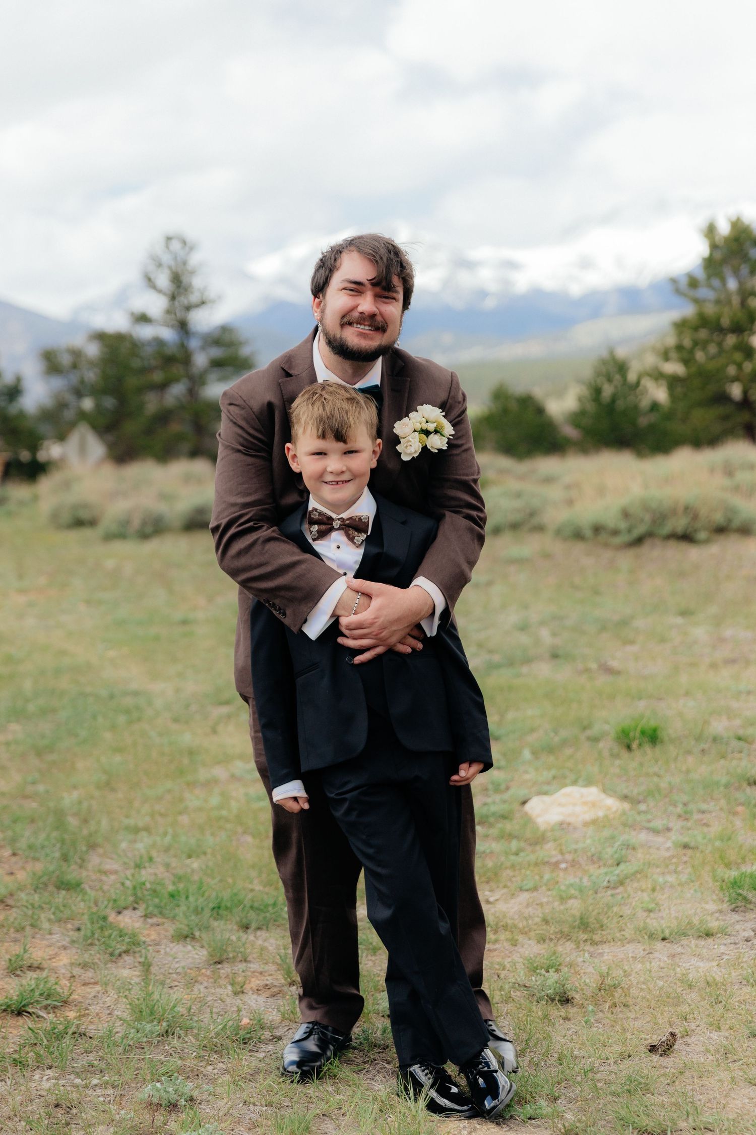 Groom in brown suit hugging young boy in tuxedo at mountain wedding outdoor setting.