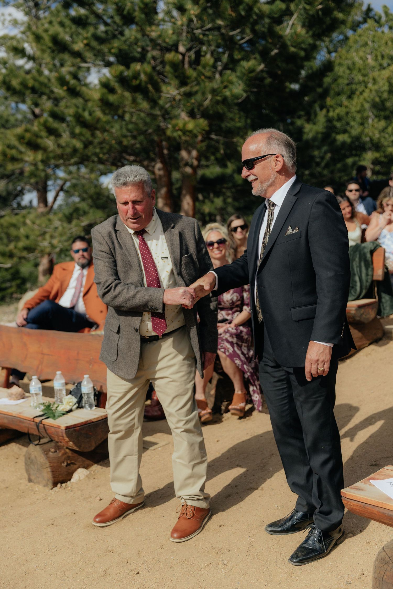 Two men in suits shaking hands at an outdoor wedding ceremony surrounded by pine trees and seated guests.