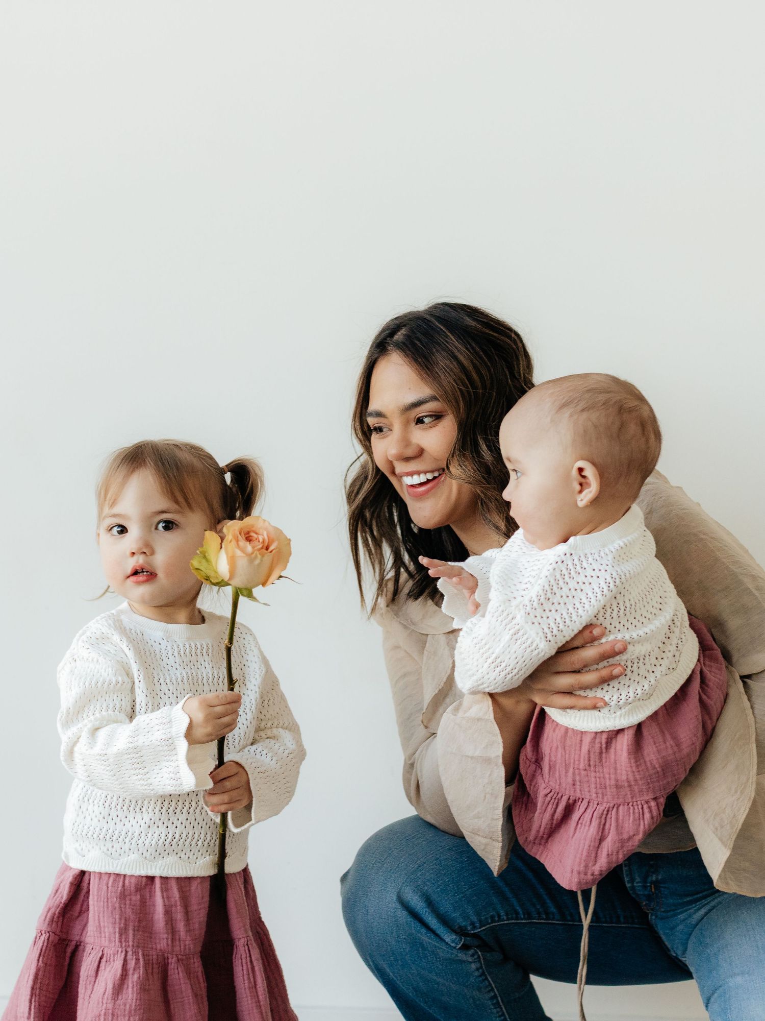 A smiling mother holds her baby girl while her toddler daughter in a pink skirt holds a peach rose.
