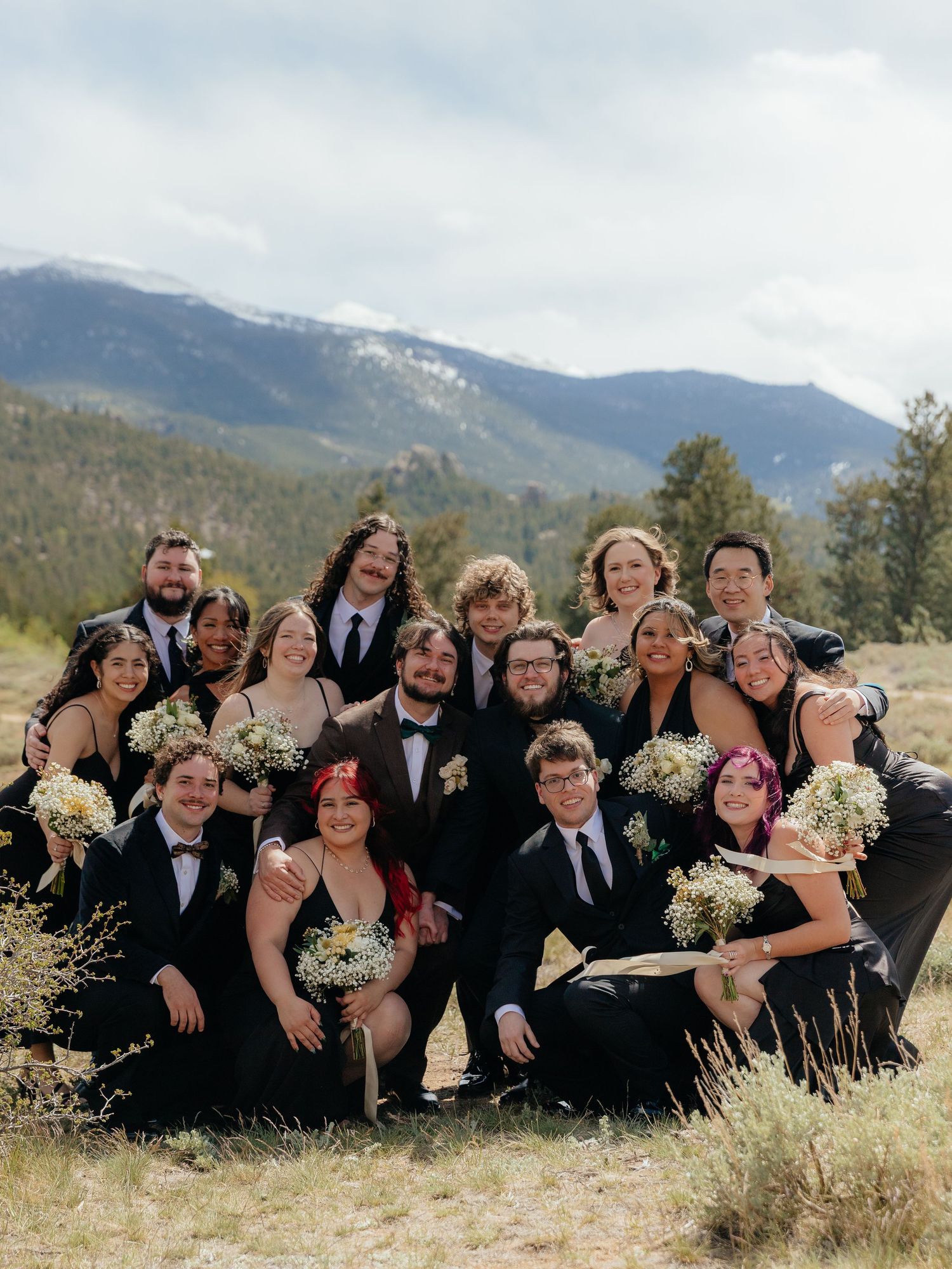 Wedding party in black attire posing outdoors with stunning Rocky Mountain backdrop and wildflower bouquets.