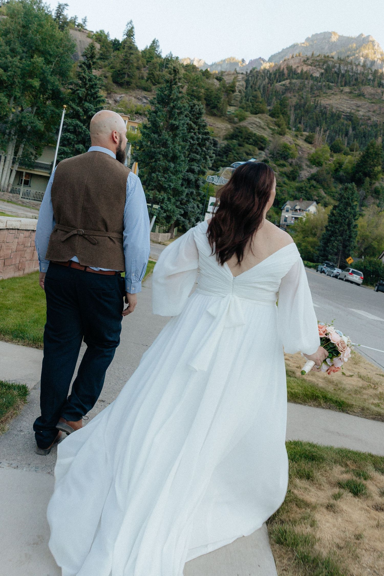 Bride and groom walking hand in hand outdoors with mountain backdrop, bride in elegant white off-shoulder gown holding bouquet.