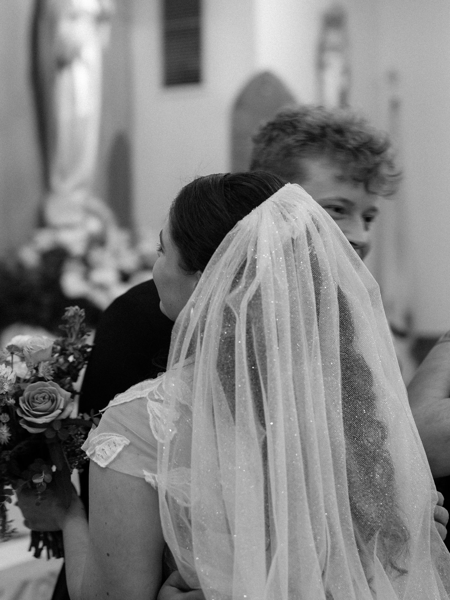 Bride wearing lace veil holds floral bouquet during church wedding ceremony in black and white photo.