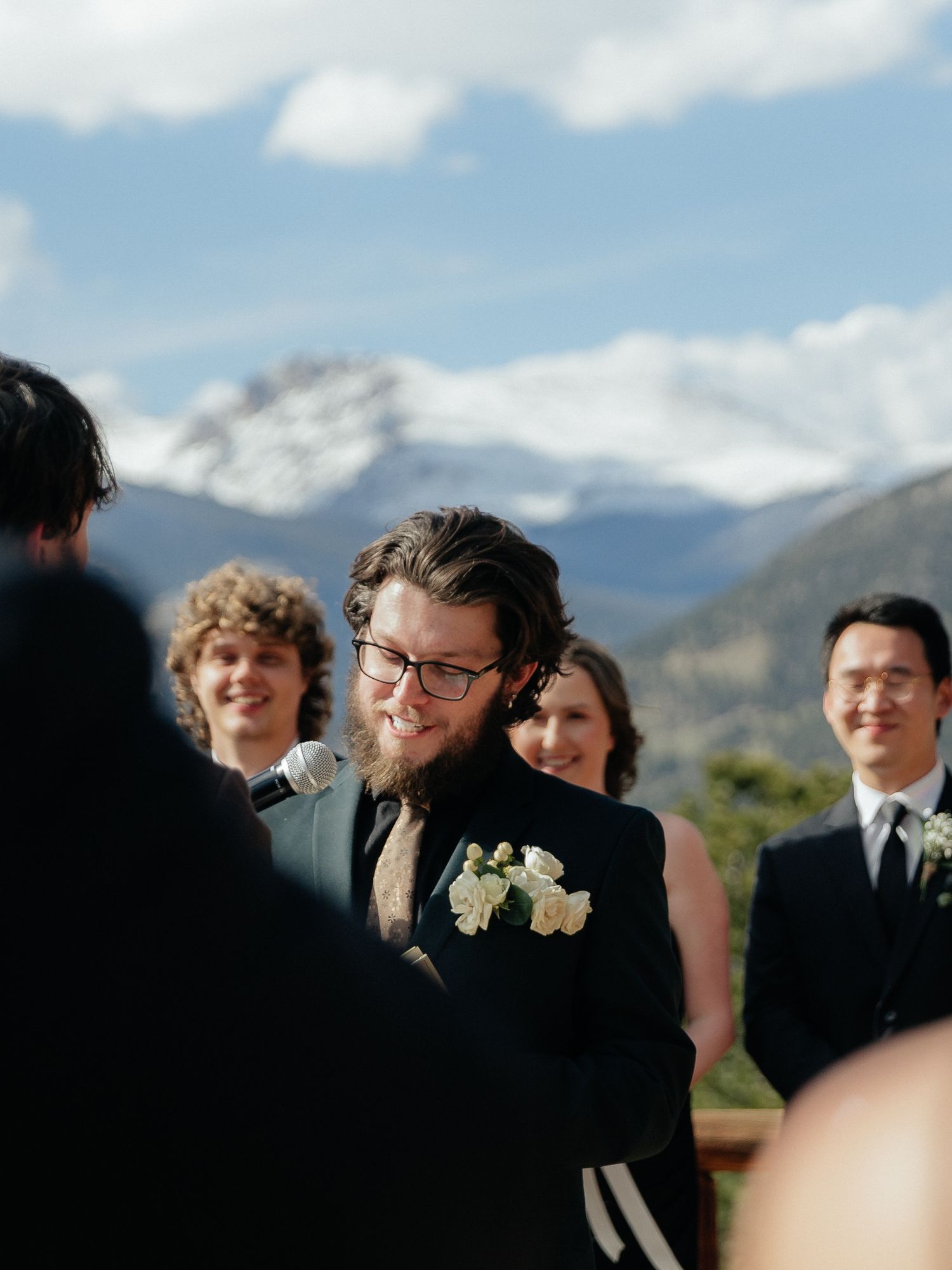 Groom with beard and glasses smiling at outdoor mountain wedding ceremony with snowy peaks backdrop.