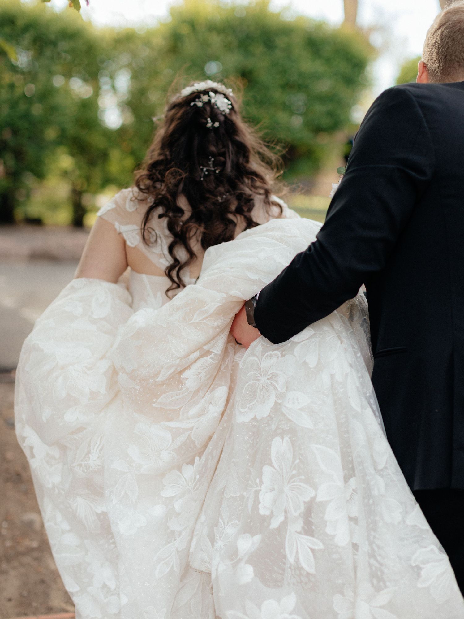 Bride in floral lace wedding gown with curly hair walks with groom in black suit outdoors.