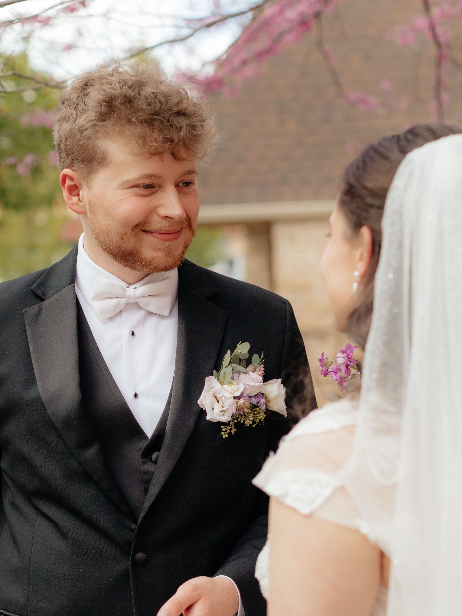 Groom in black tuxedo with white bow tie smiles at bride during outdoor wedding ceremony in spring.