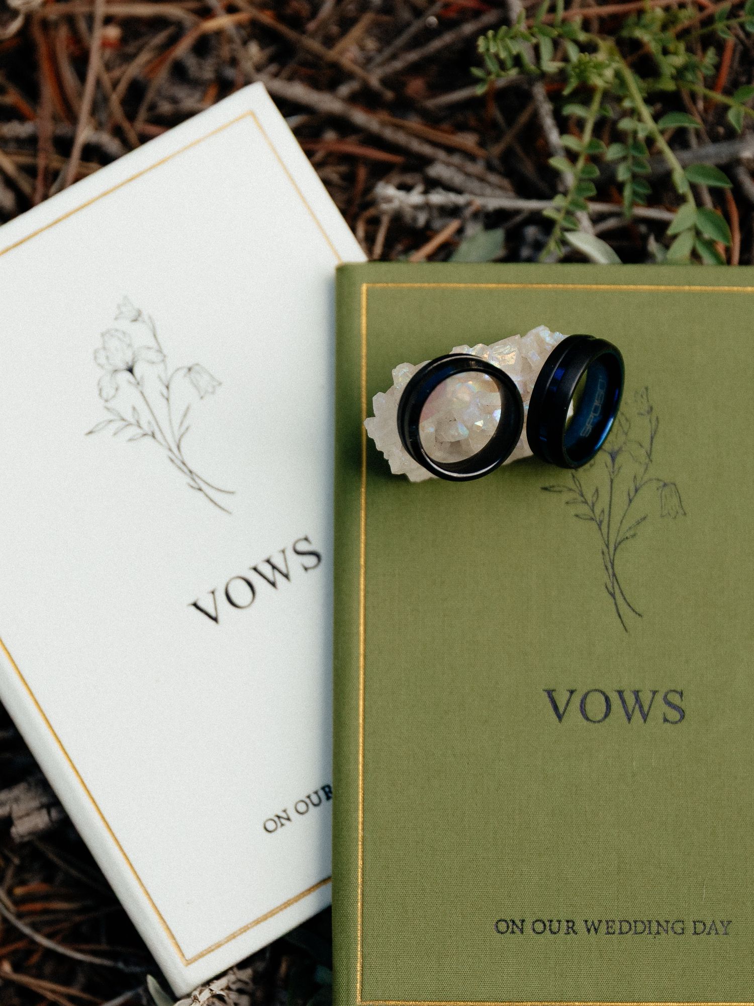 Two wedding rings rest on green and white vow books labeled Vows On Our Wedding Day.