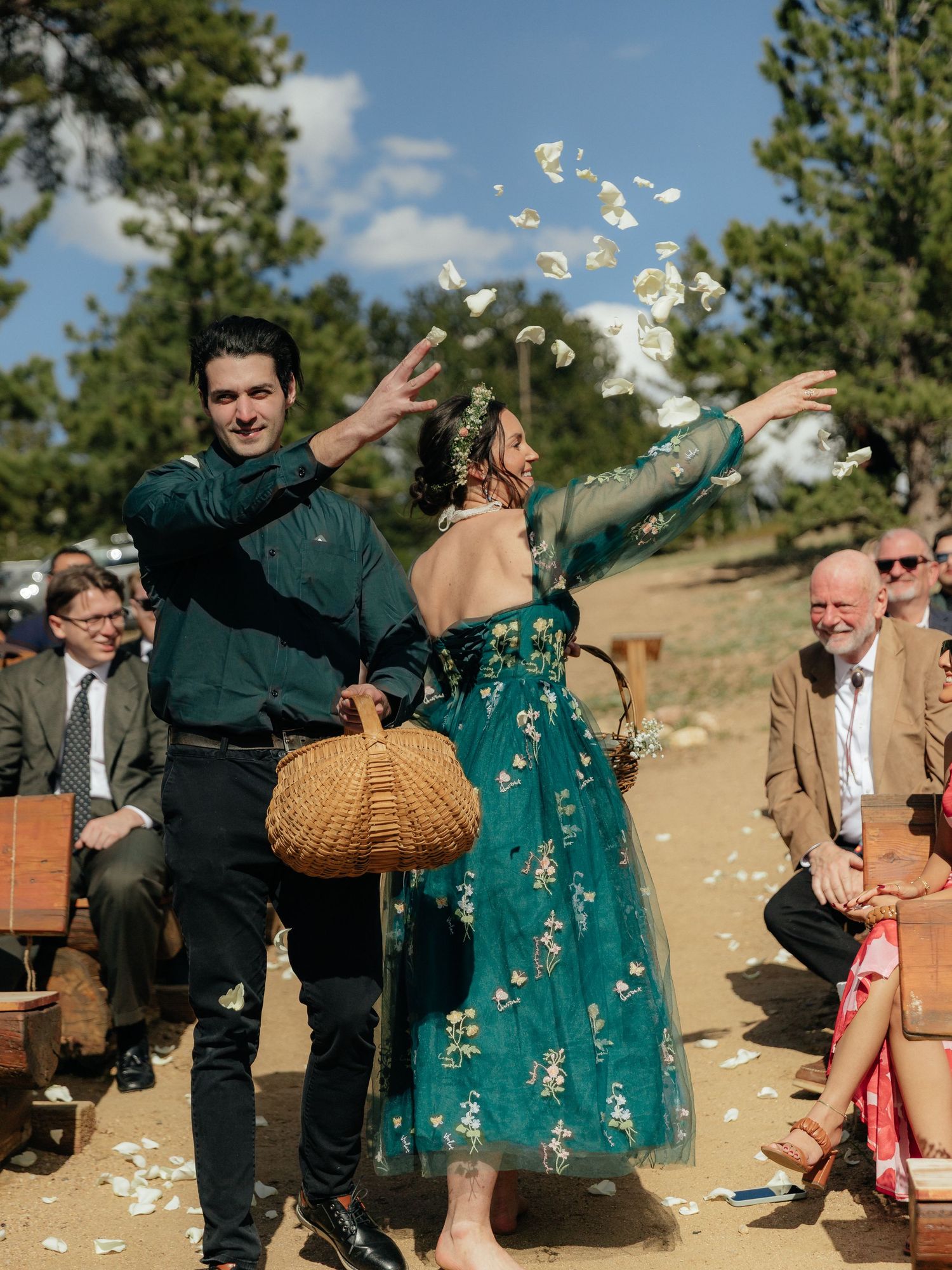 Couple tossing white flower petals at an outdoor wedding ceremony in matching teal outfits among seated guests.