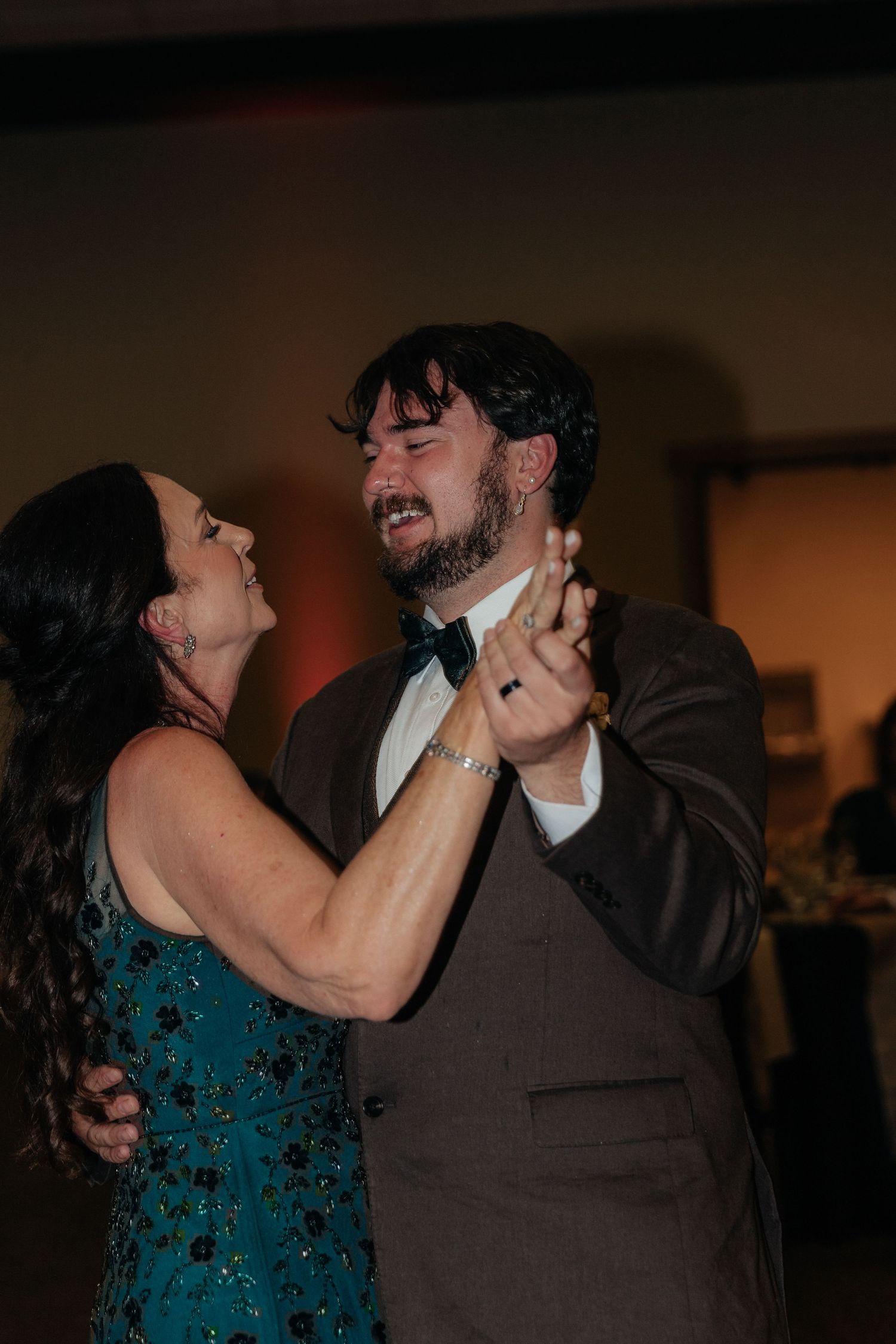 A couple shares a joyful dance, the man in a gray suit with bow tie and woman in a teal gown, smiling at each other.