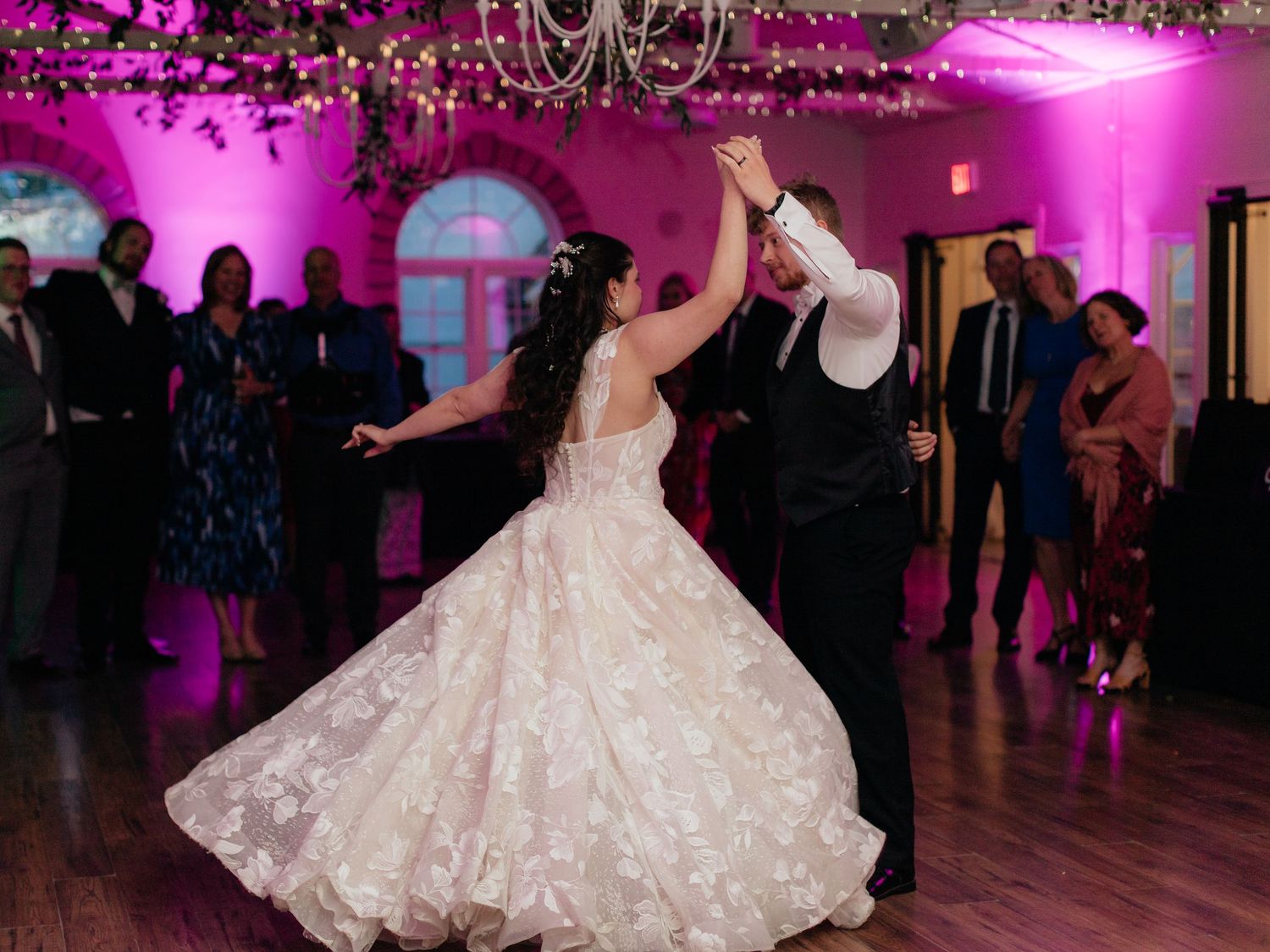 Bride in floral ballgown twirls with groom during first dance at wedding reception with pink lighting.