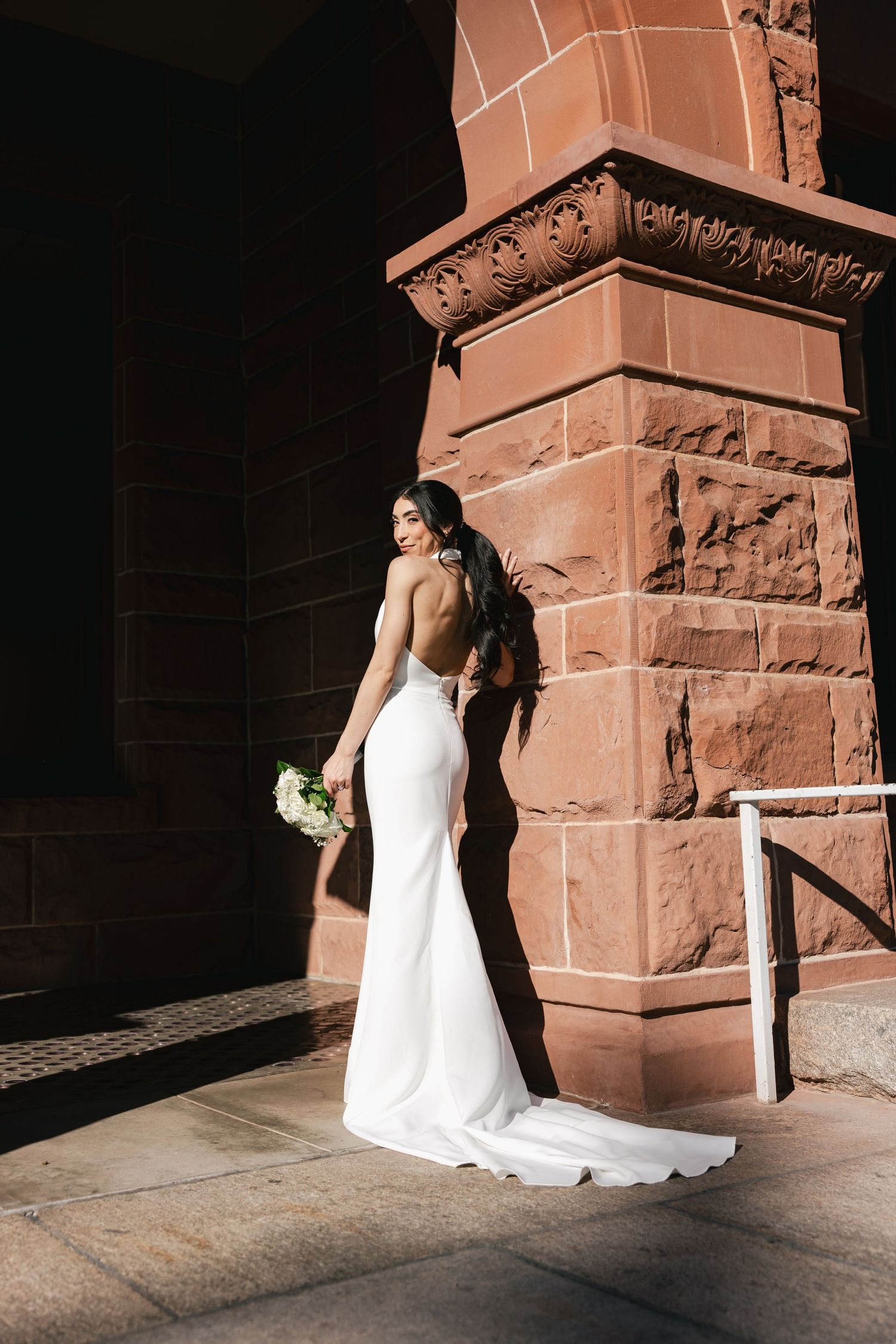 A bride in a backless white wedding gown holds a bouquet while standing against a sandstone church pillar.