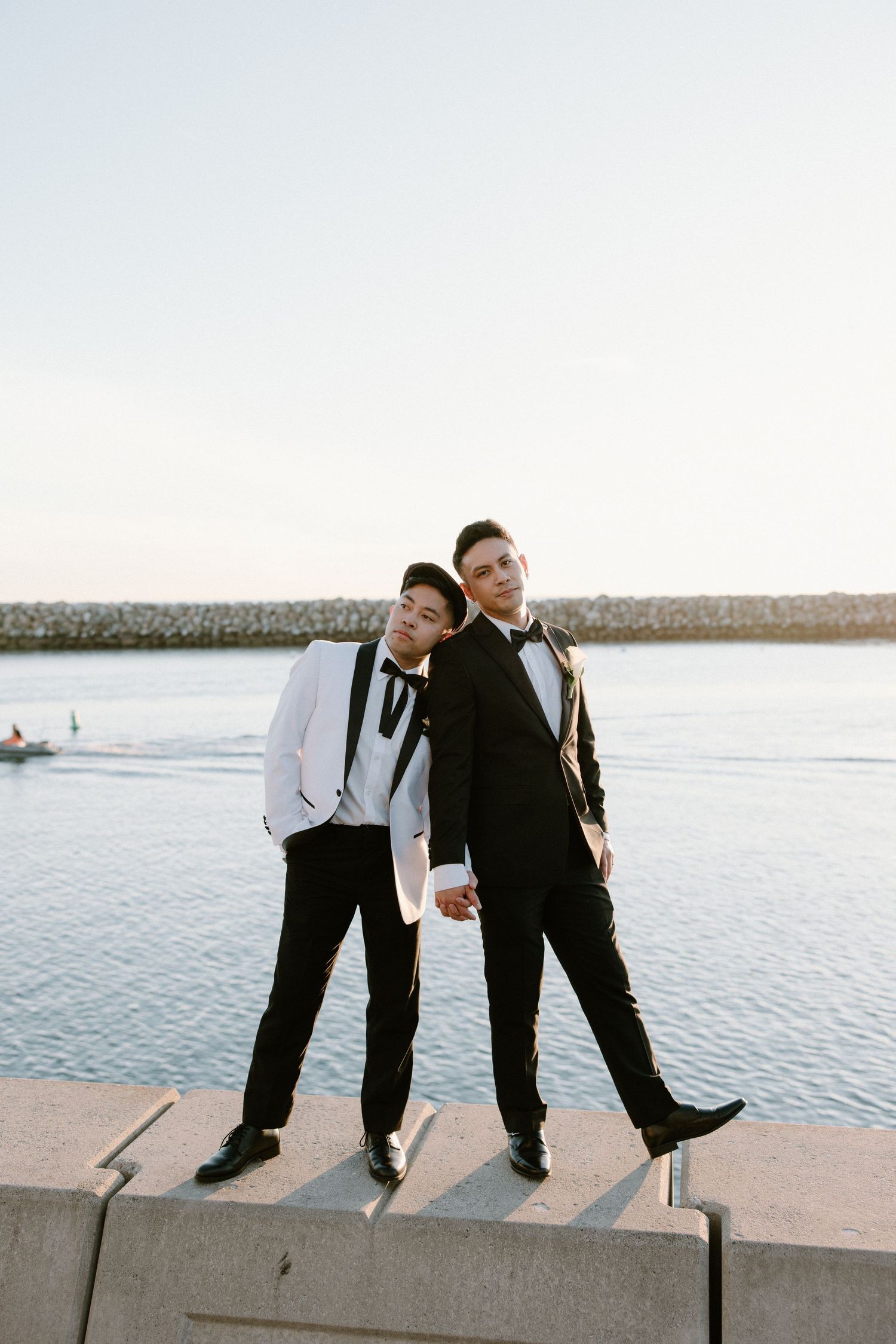 Two people in formal attire playfully lean against each other by the waterfront at sunset.