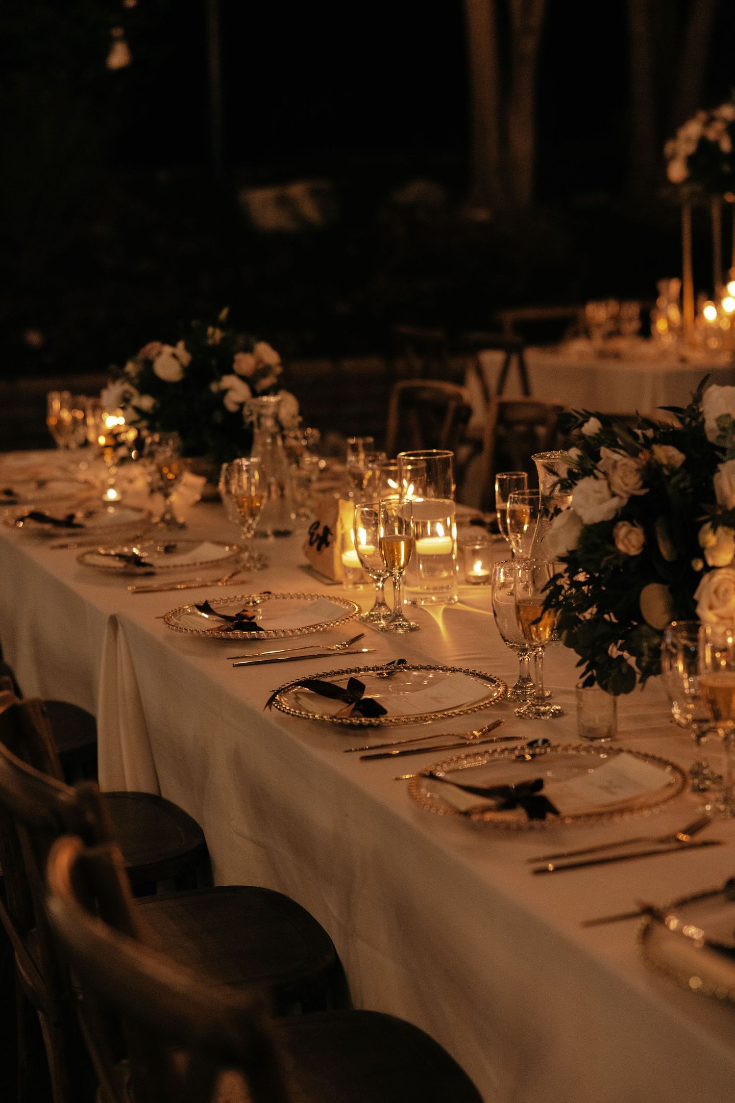 Elegant candlelit wedding reception table with white linens, floral centerpieces and formal place settings in warm lighting.