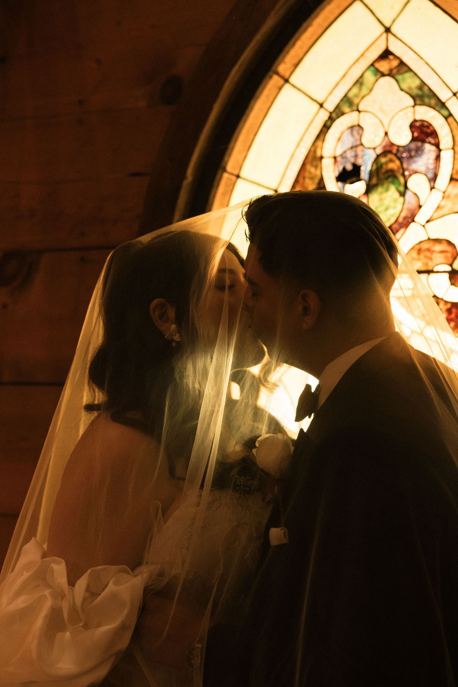Bride and groom sharing a kiss under a veil in a church, backlit by a golden stained glass window.