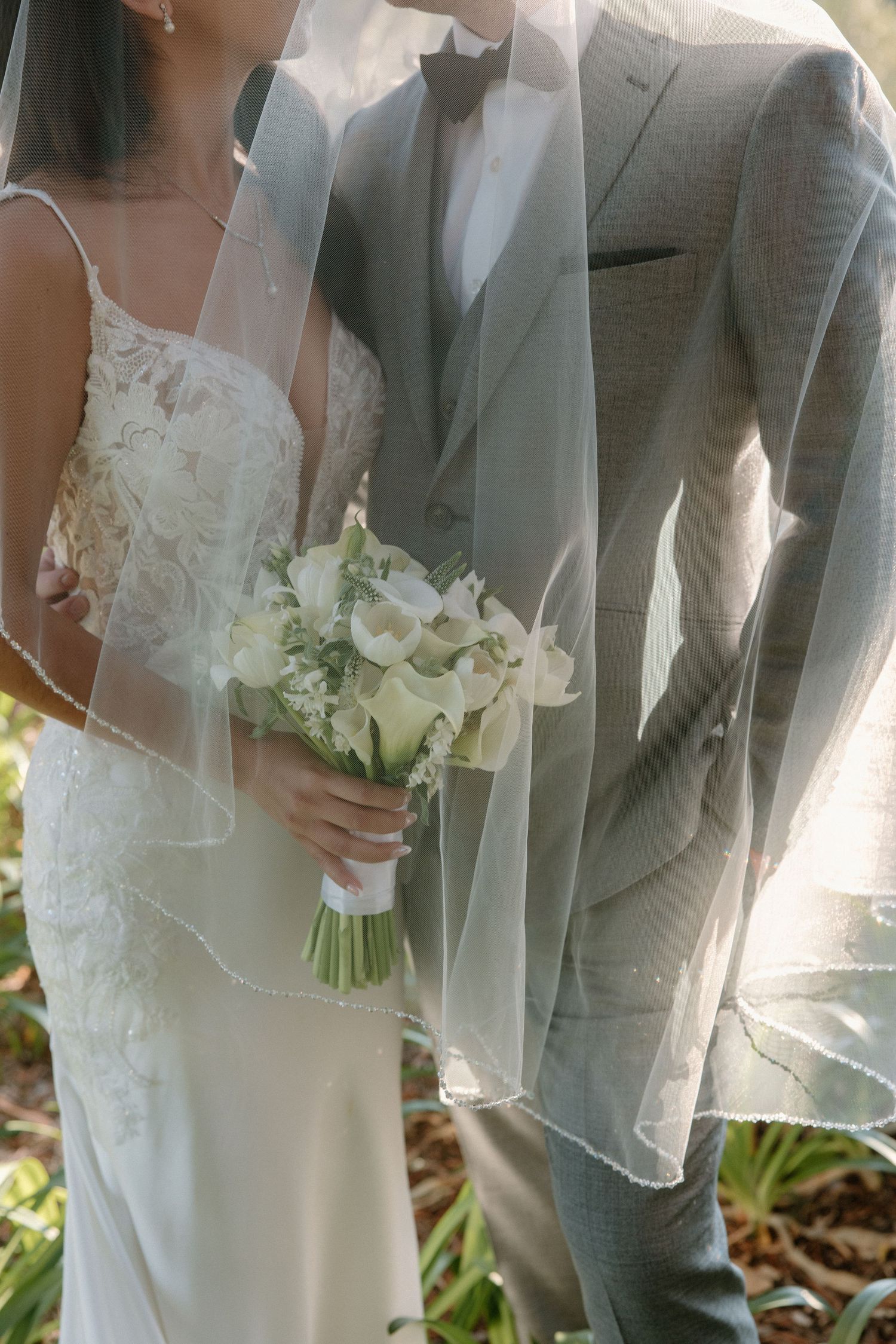 Close-up details of a bride and groom at a wedding ceremony showing white dress, gray suit, and white floral bouquet.