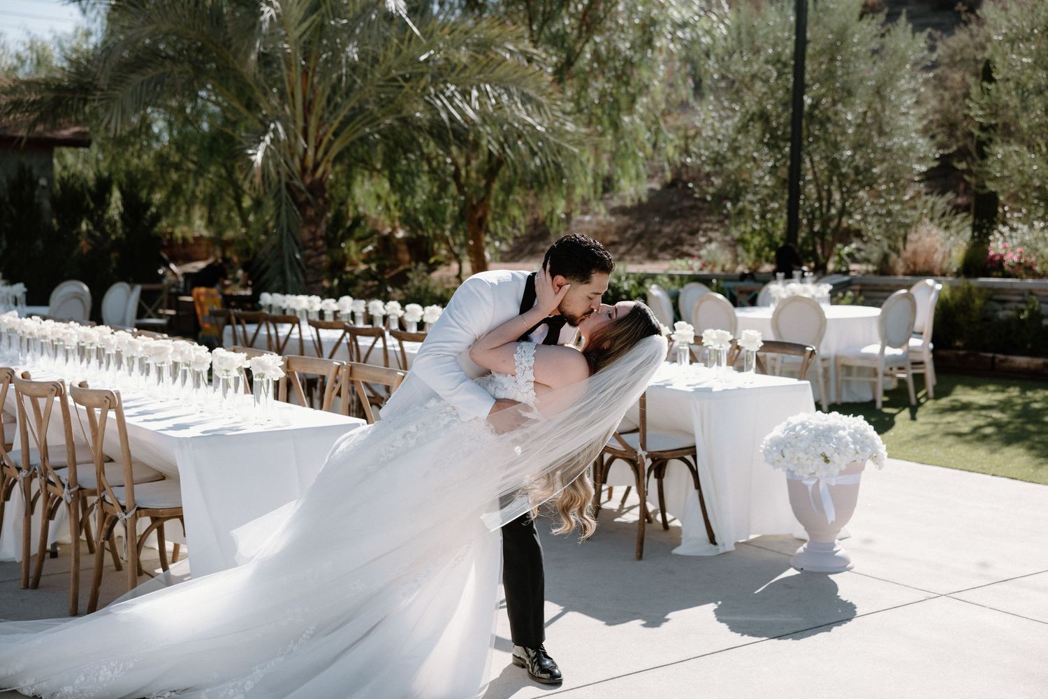 An elegant outdoor wedding reception setup with wooden chairs and white linens under a canopy of trees.