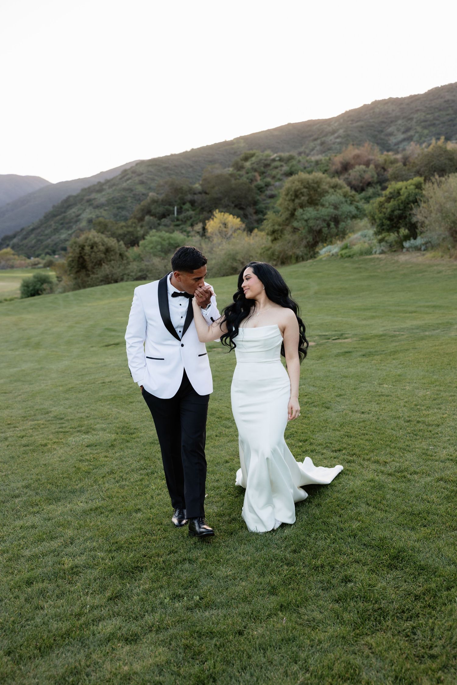 A groom in a white tuxedo kisses his bride's hand as she smiles in a mint green gown on a lush hillside.