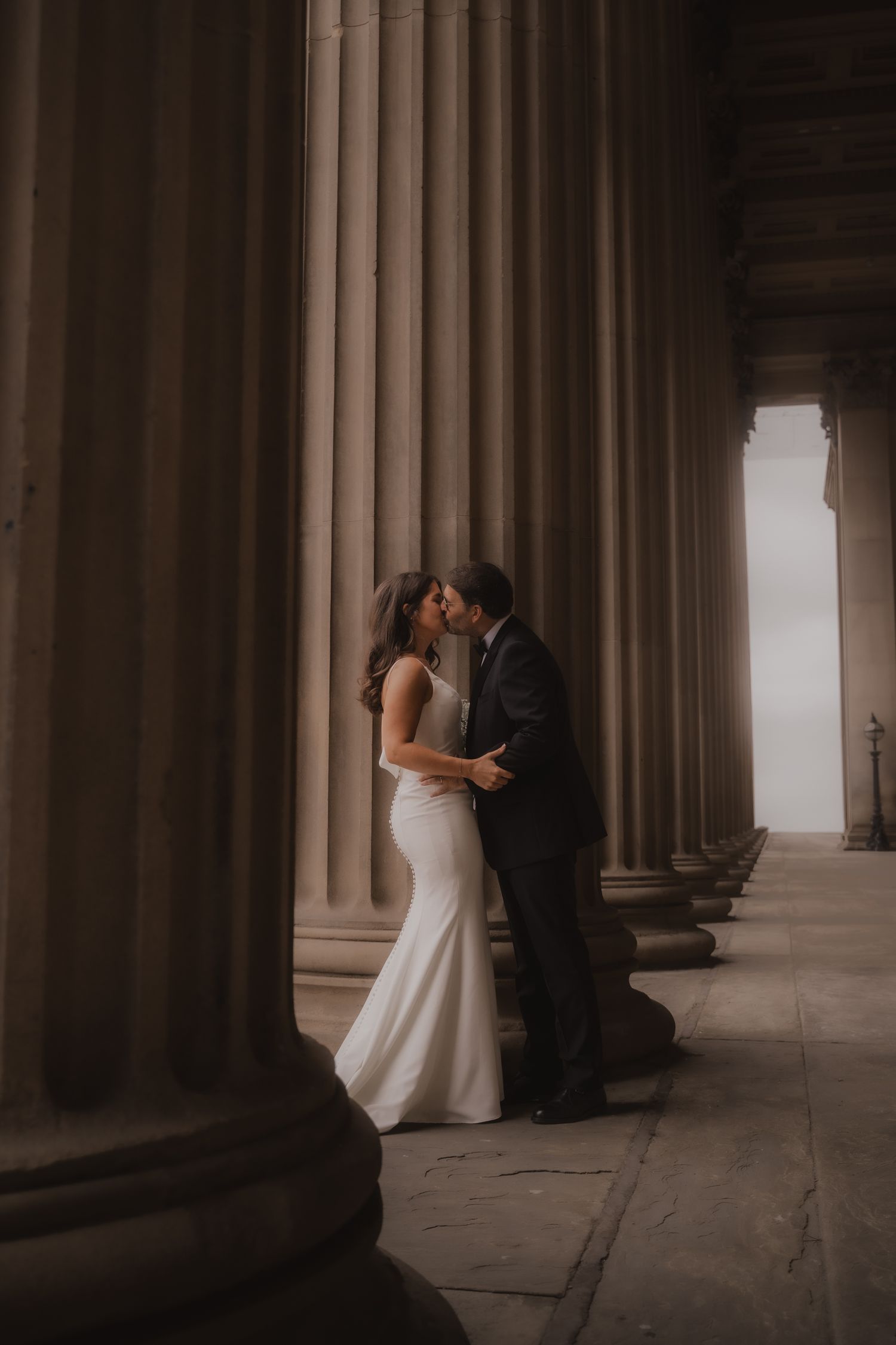 A romantic wedding couple embraces between elegant stone columns in a moody, atmospheric architectural setting.
