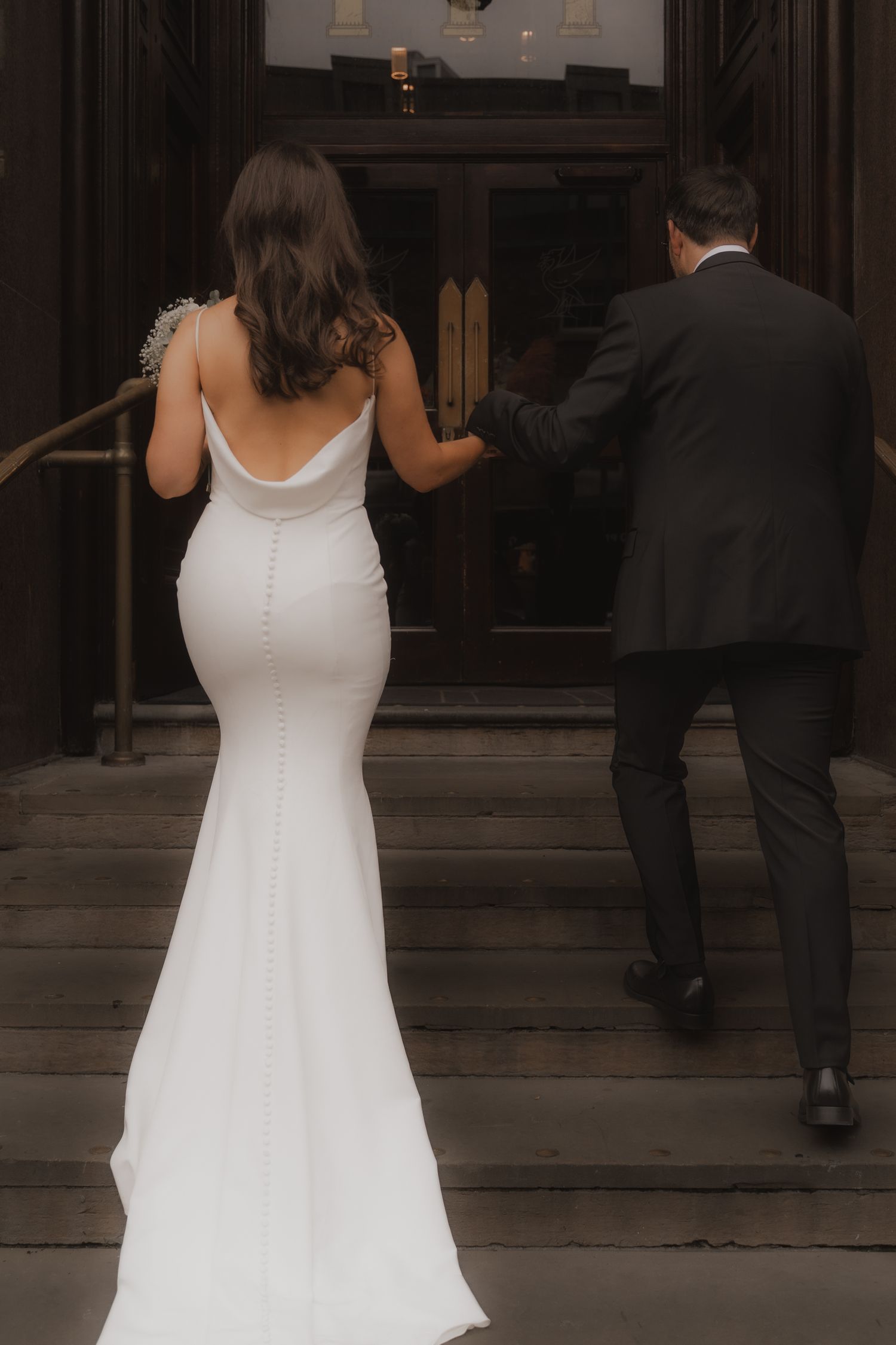A couple in formal attire walks into a building, with the person in white wearing a flowing dress.