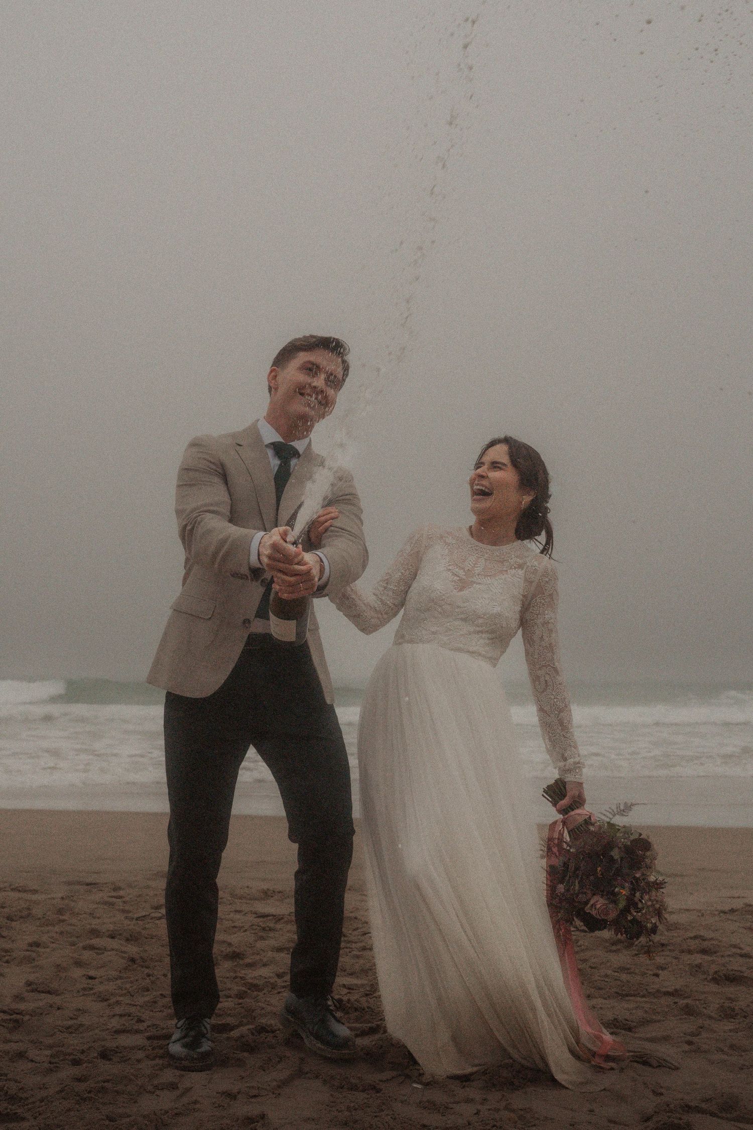Newlyweds share a champagne moment on misty beach.