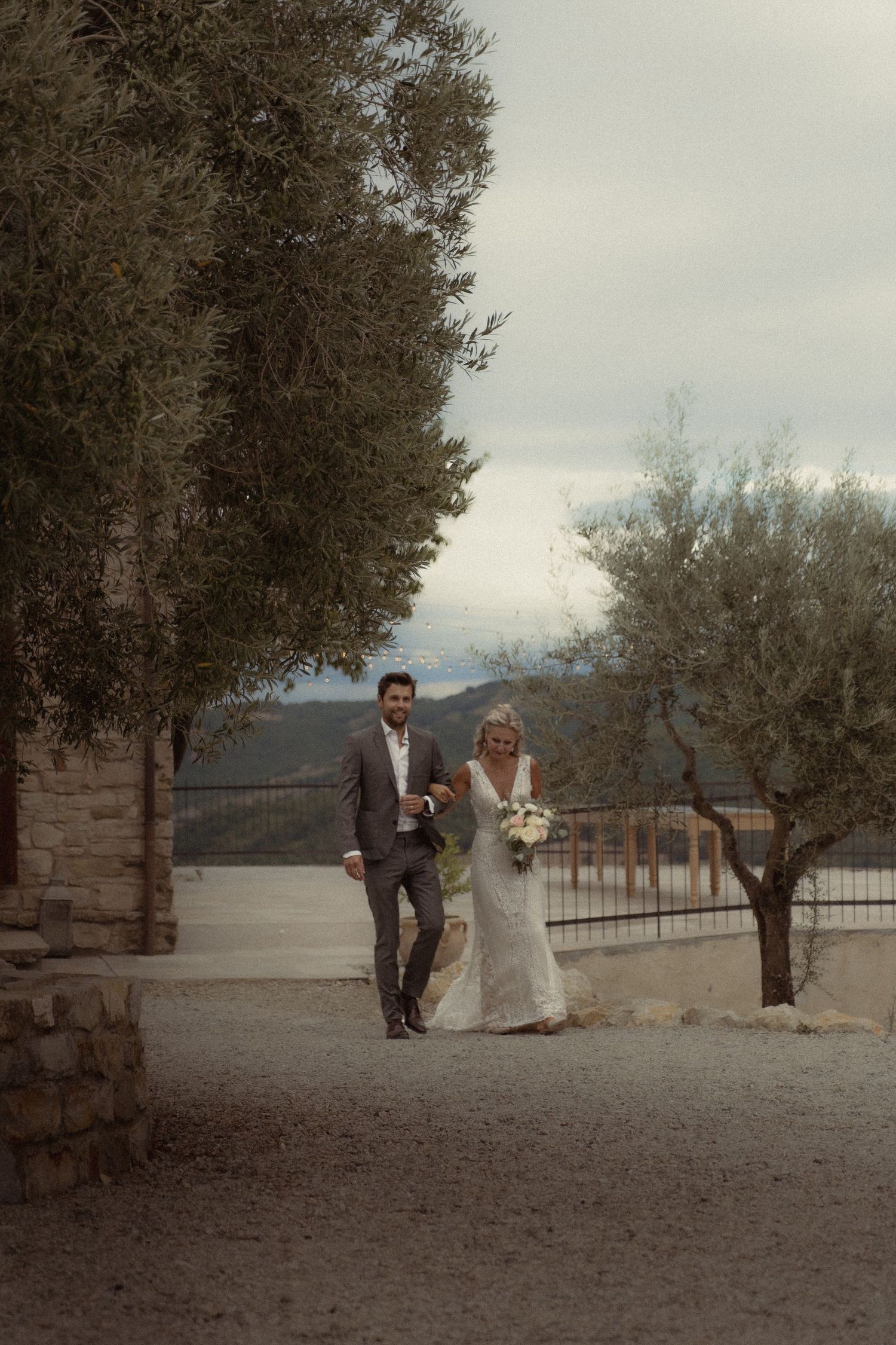 A couple in wedding attire strolls through an elegant stone pathway with mountain views.