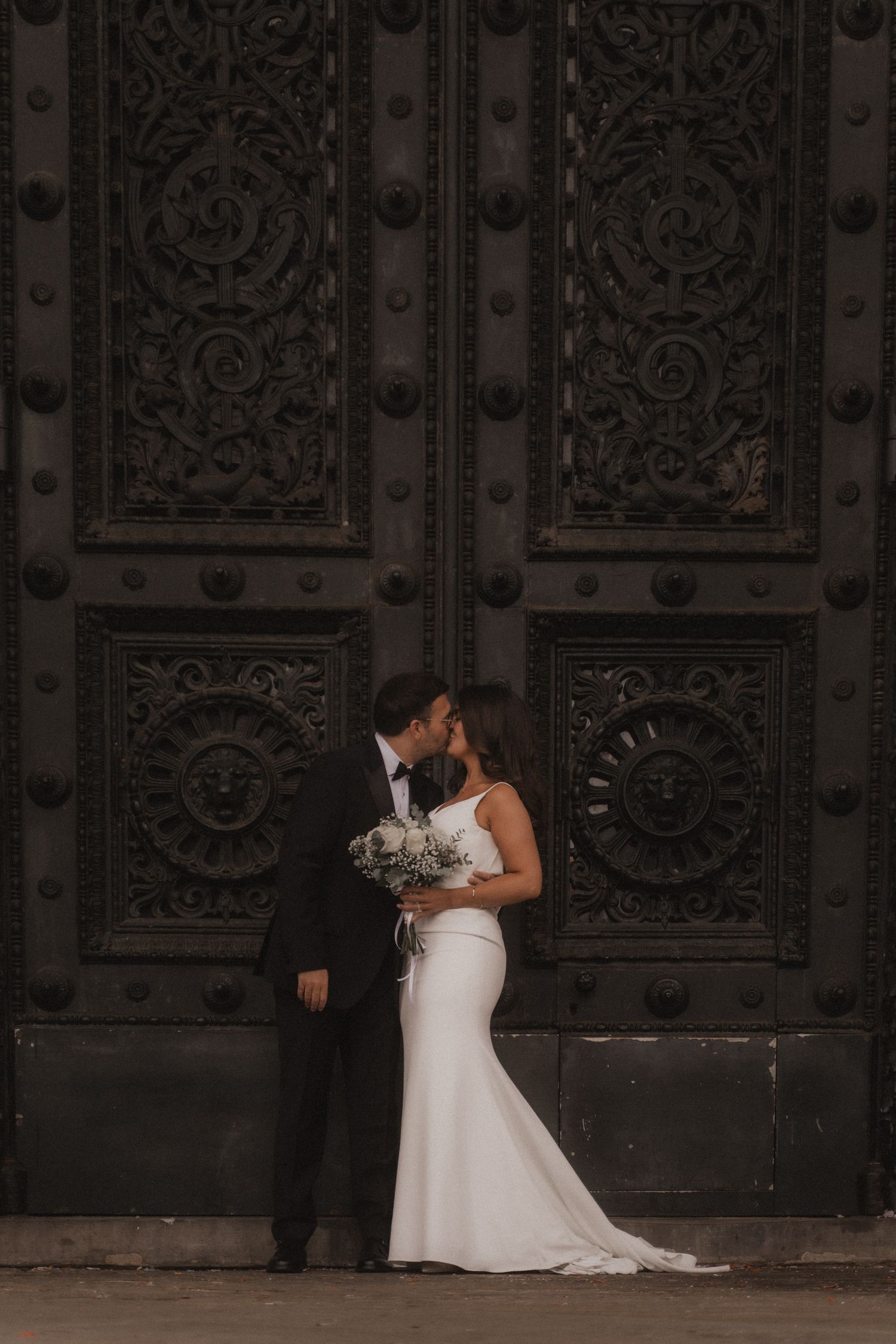 A bride in a white wedding dress sits thoughtfully on stone steps beside ornate vintage metal doors.