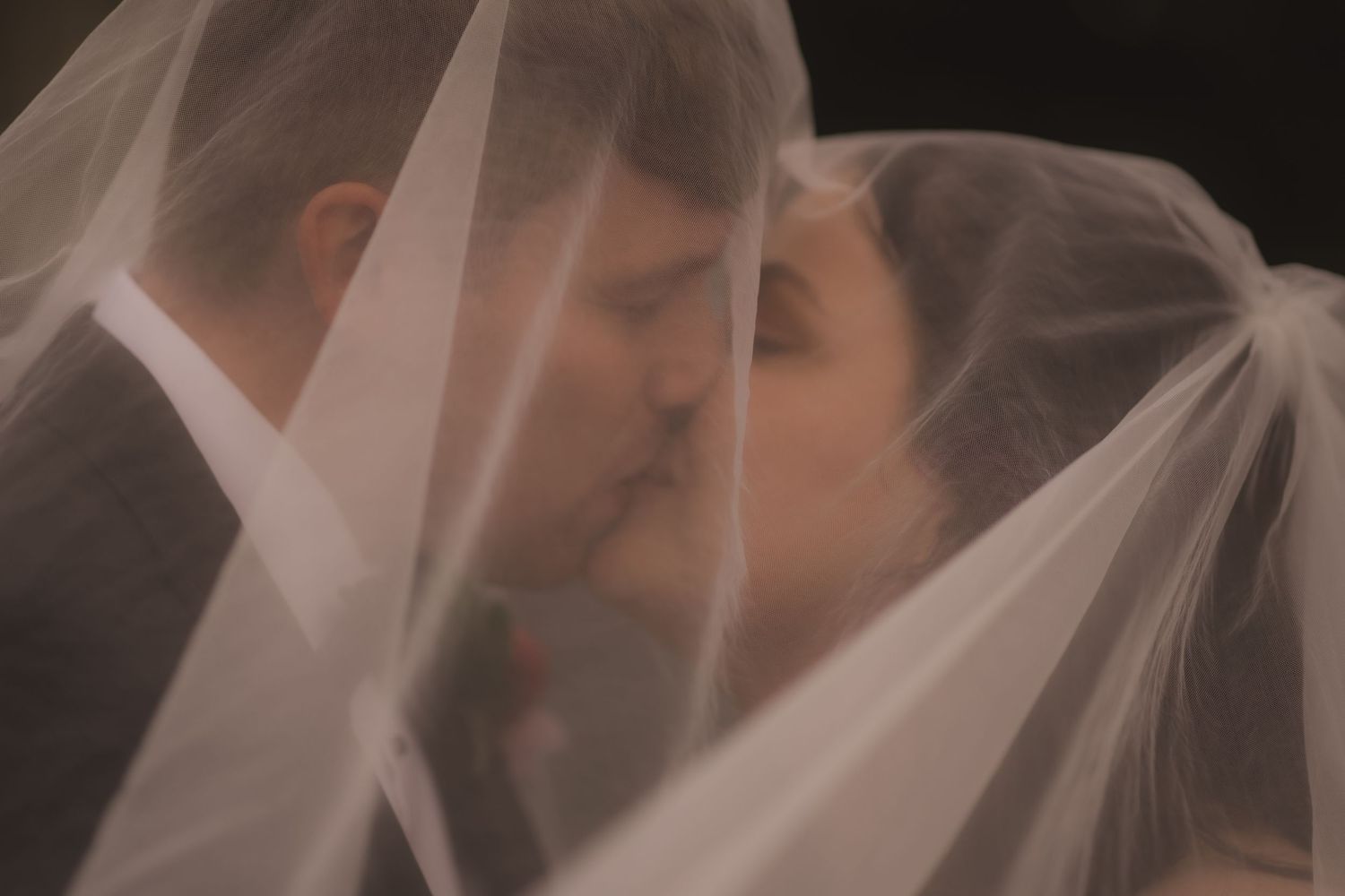 A romantic couple shares an intimate kiss under a sheer white veil during their wedding ceremony.