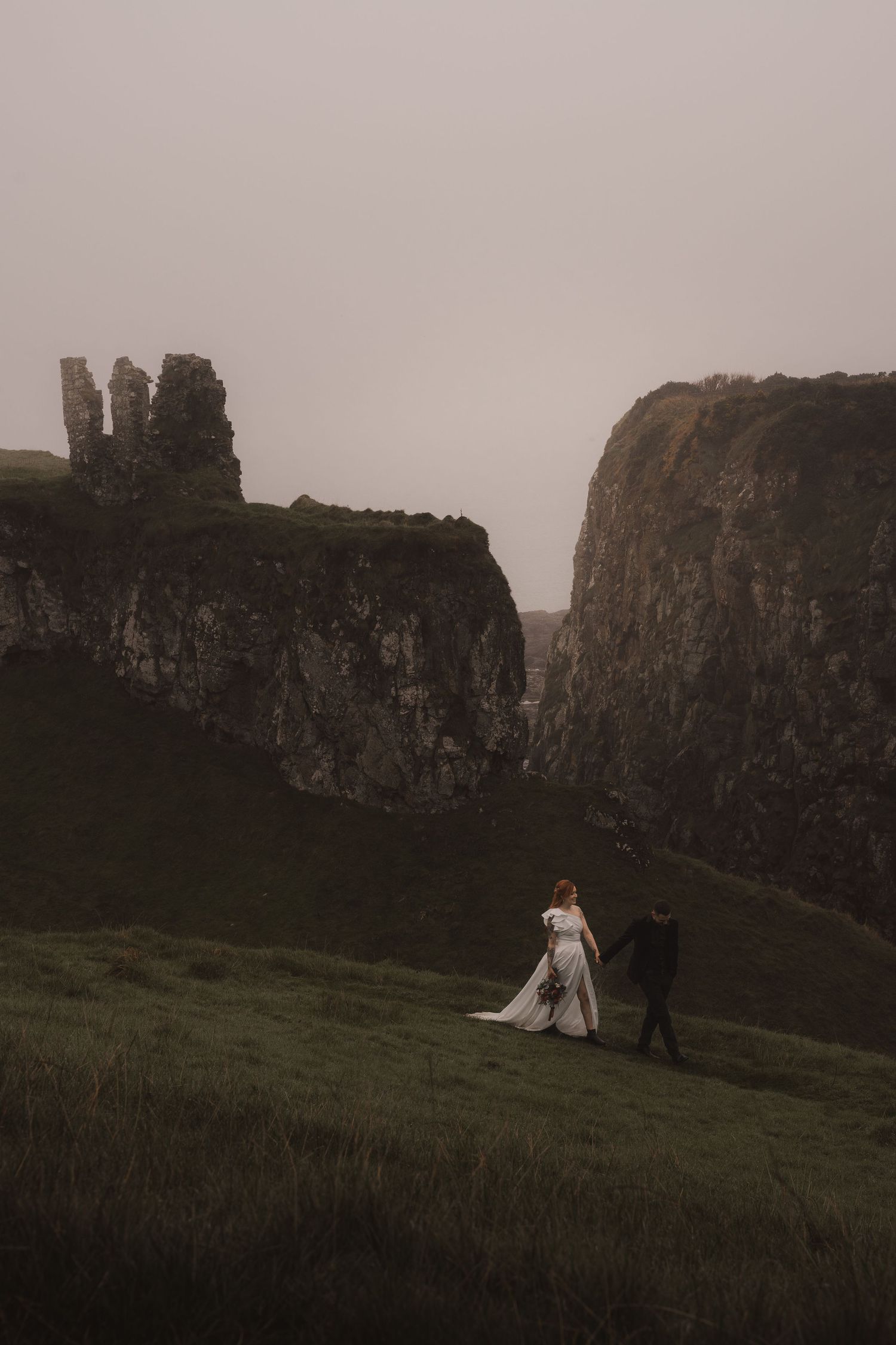 A wedding couple stands small against massive coastal cliffs in a moody landscape photograph.