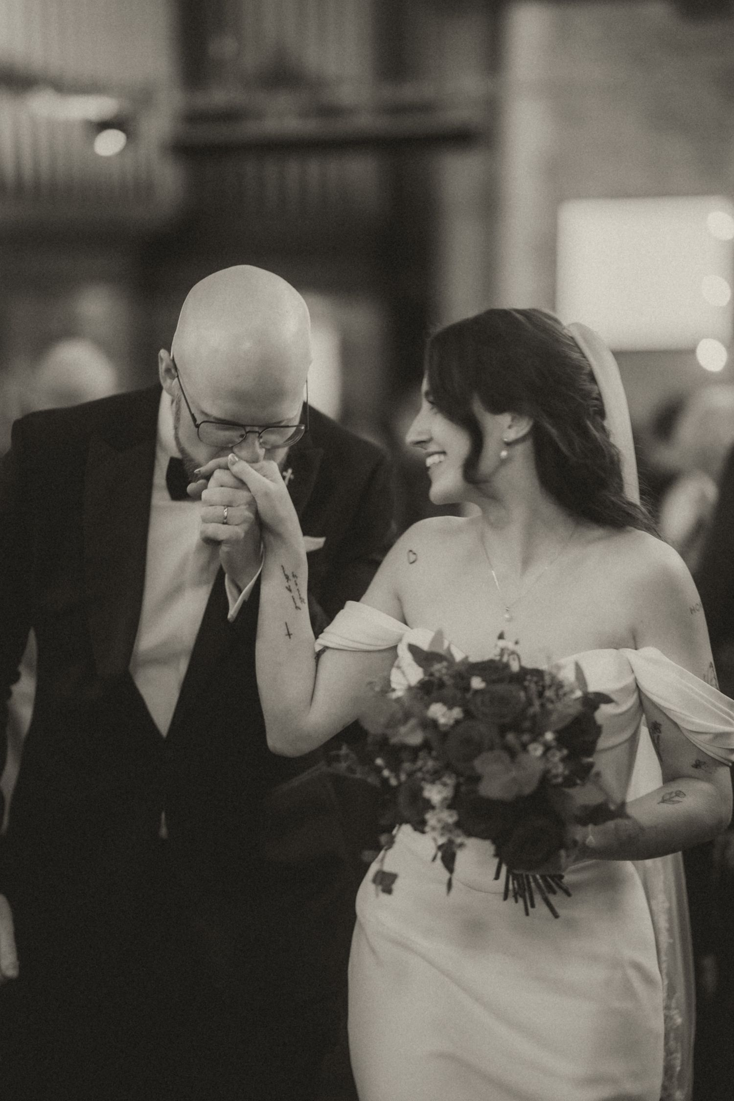 Black and white photo of emotional groom wiping tears beside smiling bride holding bouquet during wedding ceremony.