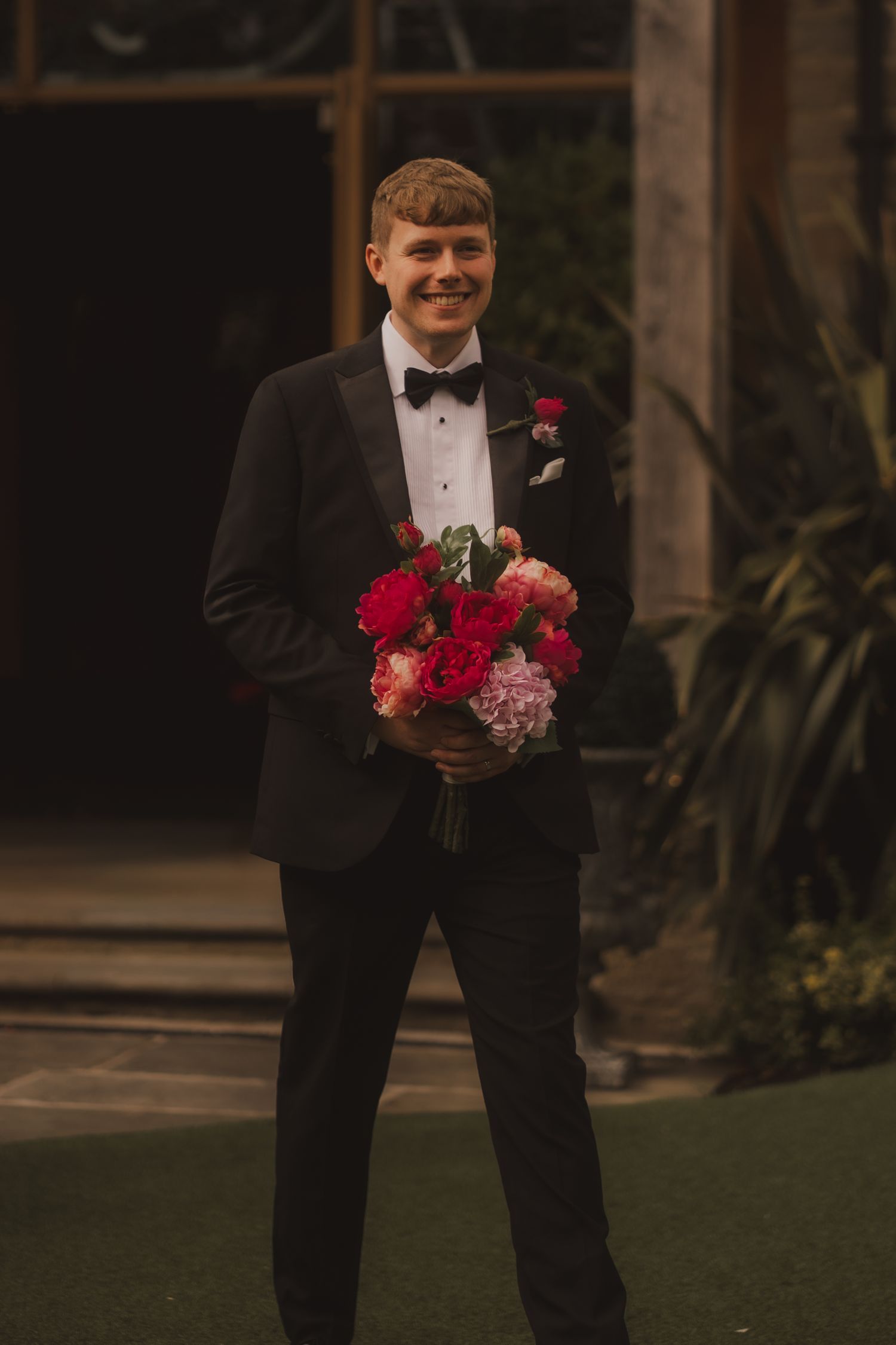 A person in a black tuxedo and bow tie holds a vibrant bouquet of red and pink flowers at an outdoor venue.