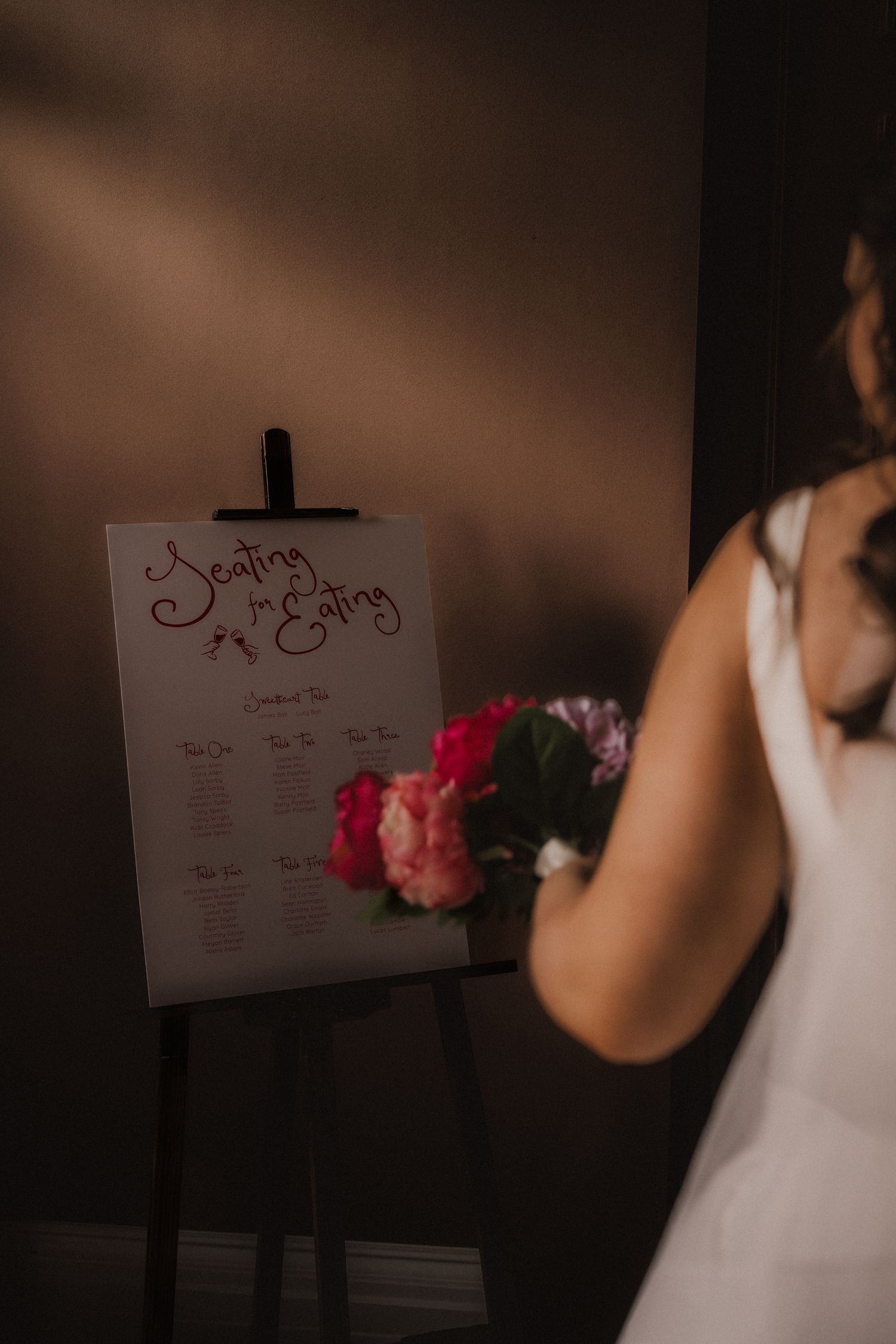 Wedding seating chart displayed on an easel with pink floral decorations in a dimly lit room.