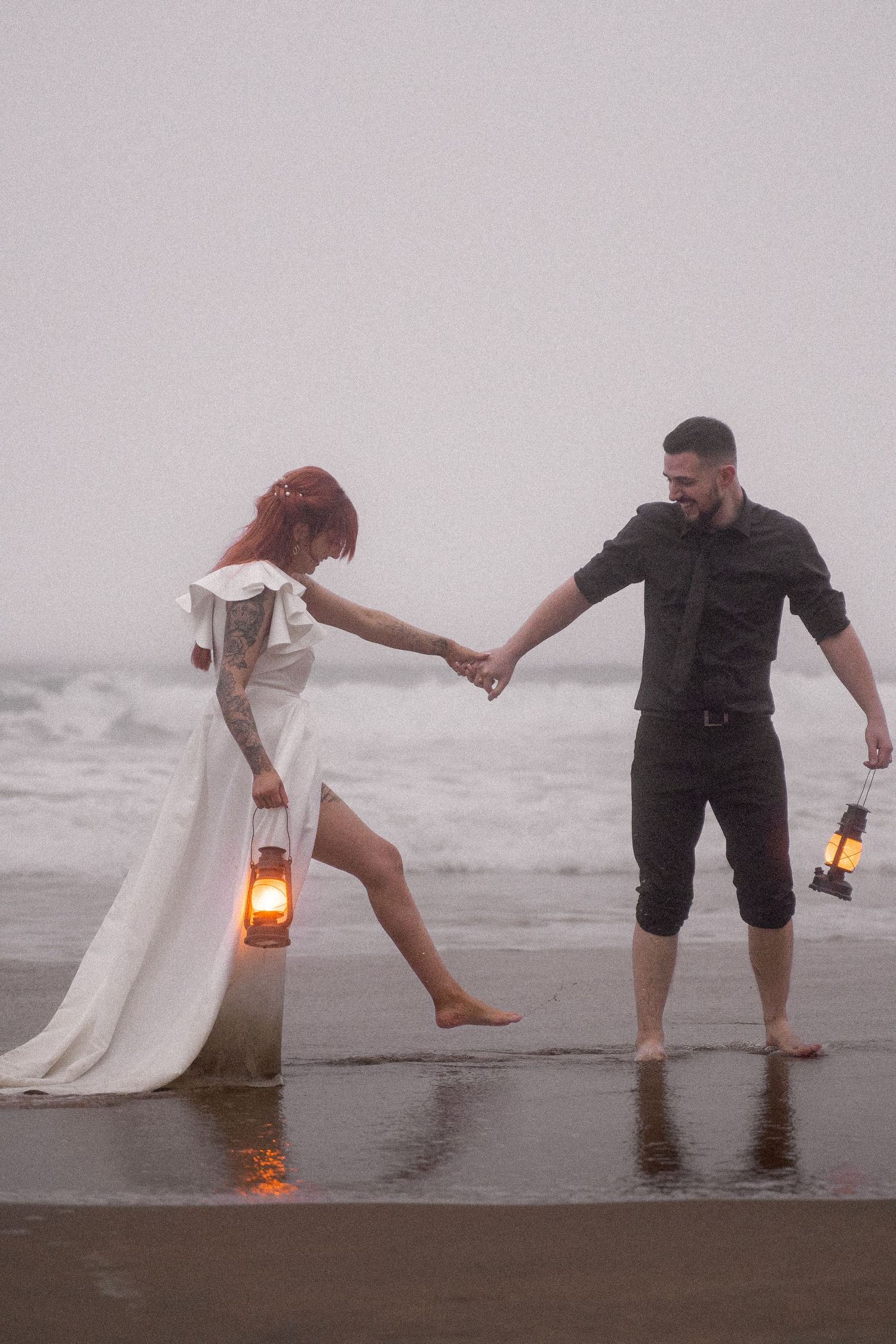 A couple holding lanterns playfully splashes in the ocean waves during their beach wedding photoshoot.