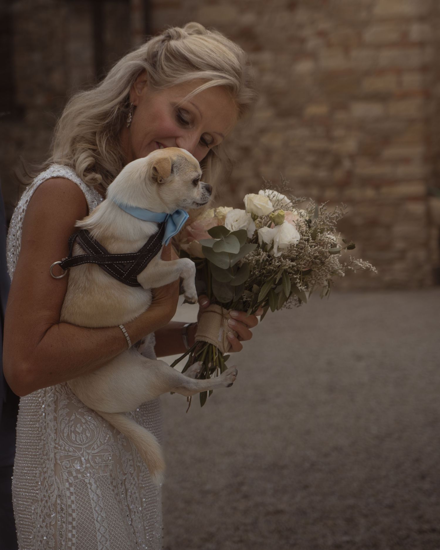 Bride in white dress holds a cat and bouquet against brick wall backdrop.