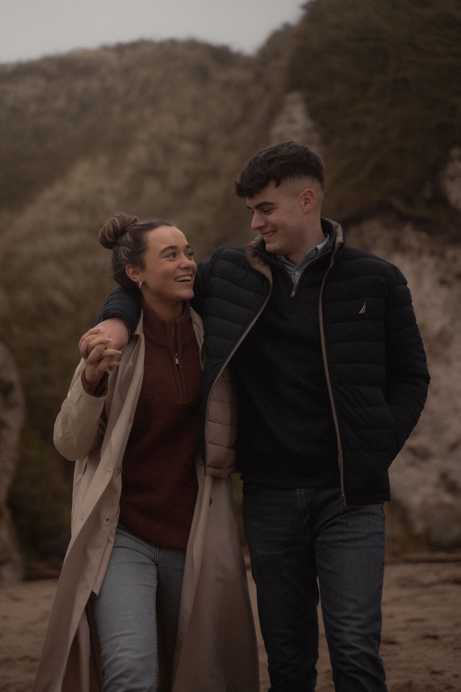 Two people walking and laughing outdoors in autumn attire with mountains in the background.