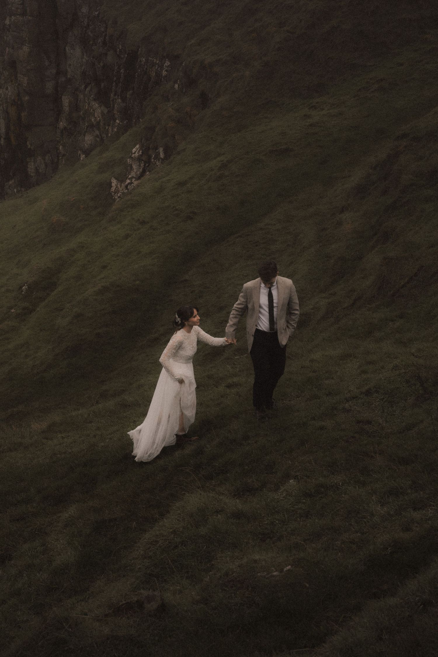 Wedding couple walking hand in hand on a steep grassy hillside.