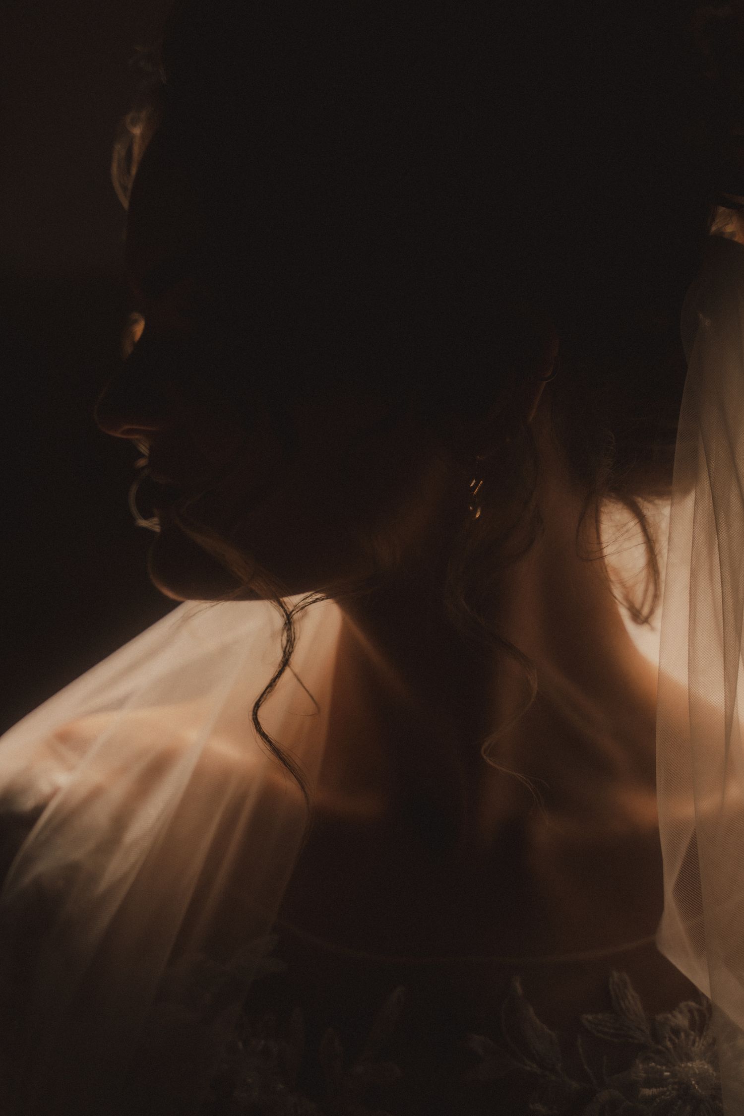 A bride in soft golden light wearing a veil and lace dress, elegant earring visible in a moody dark portrait.