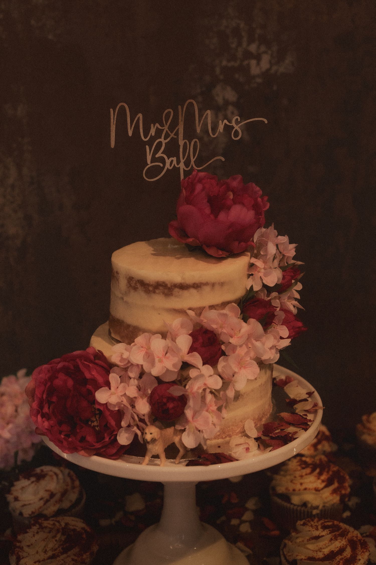 Elegant white wedding cake decorated with pink and burgundy flowers on a cake stand against a dark background.