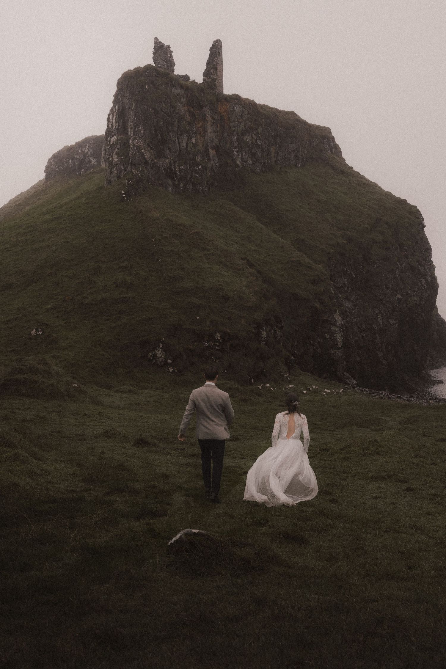 A series of moody landscape photos showing people walking up a steep, misty hillside in wedding attire.