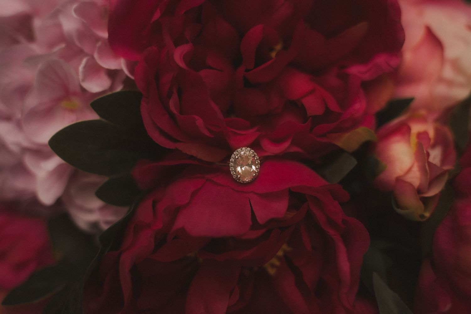 A sparkling diamond ring nestled among deep red peony petals in soft lighting.