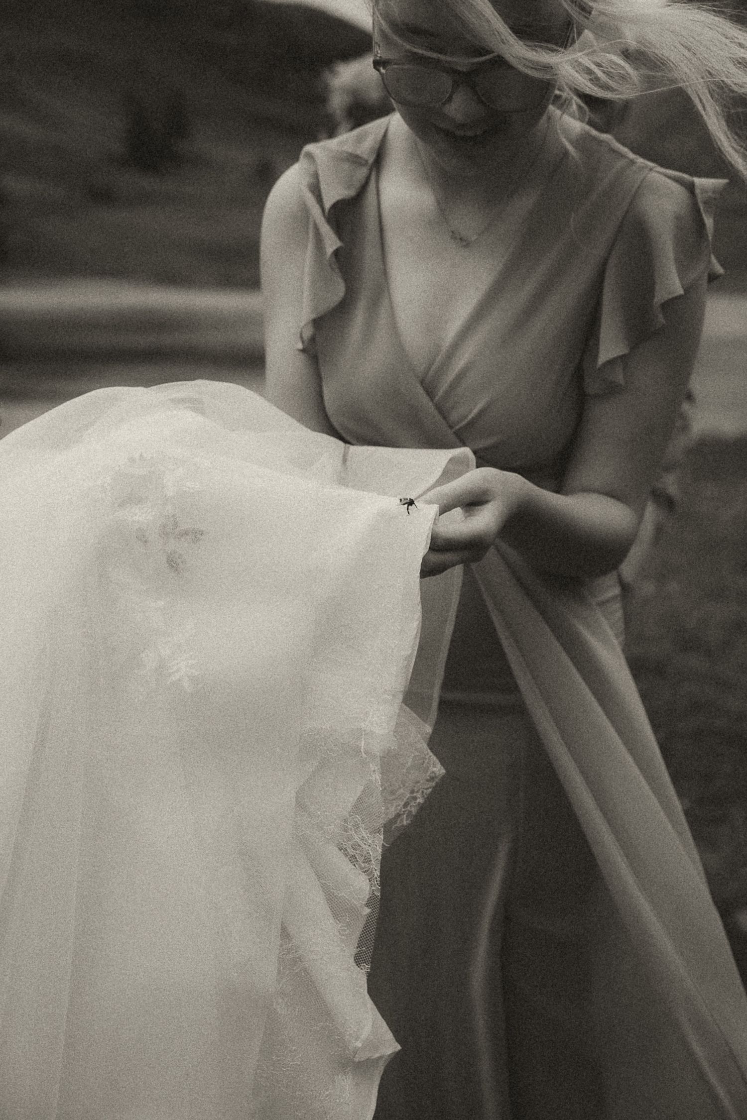Two people in elegant formal wear prepare wedding dress details at an outdoor garden venue.