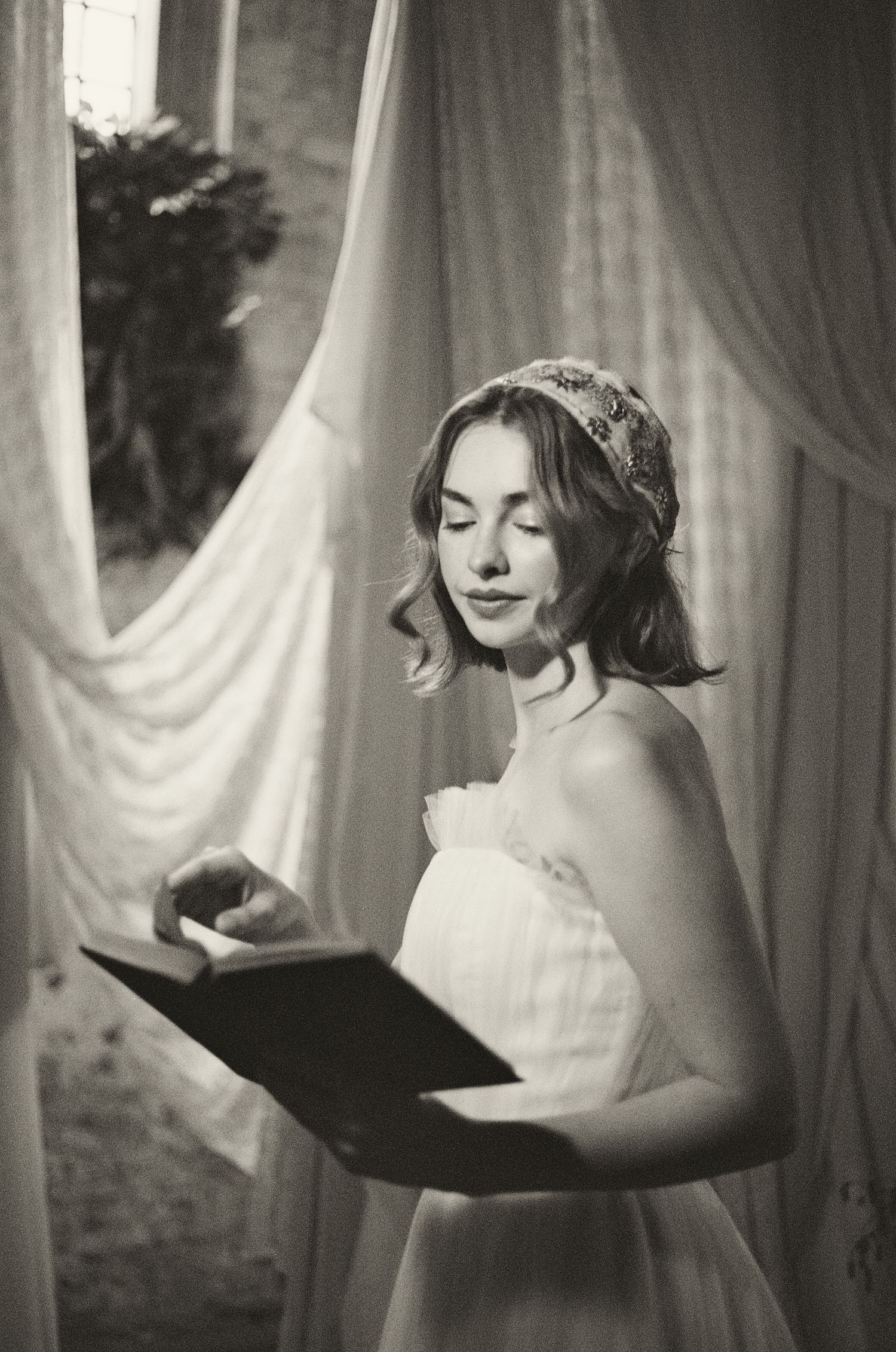 A black and white portrait of someone in a white dress reading a book near sheer curtains in soft natural light.