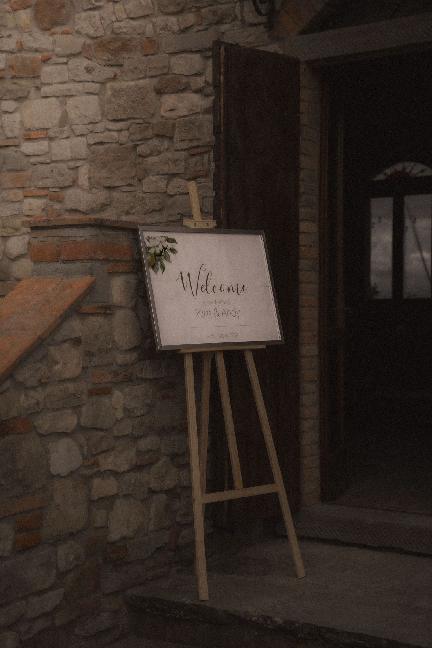 A wooden welcome sign on an easel stand placed against a rustic stone wall entrance.