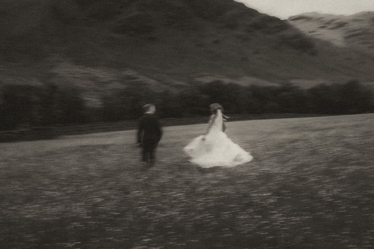 Black and white artistic photograph of two figures walking across a meadow with mountains in the background.