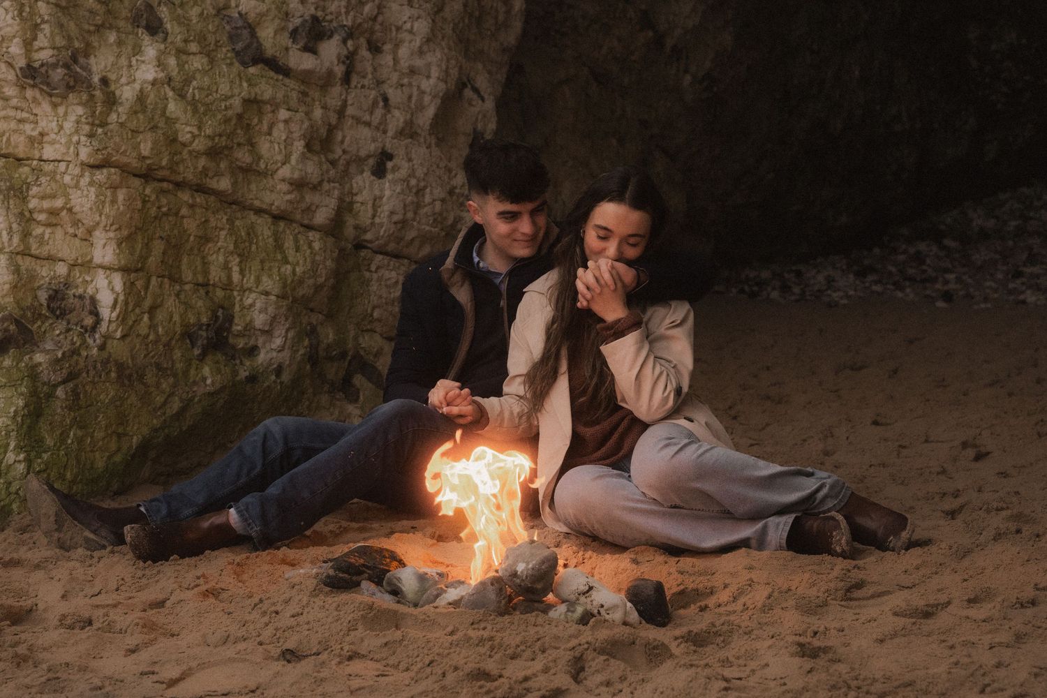 A romantic evening scene with a small campfire glowing inside a cave on a sandy beach.