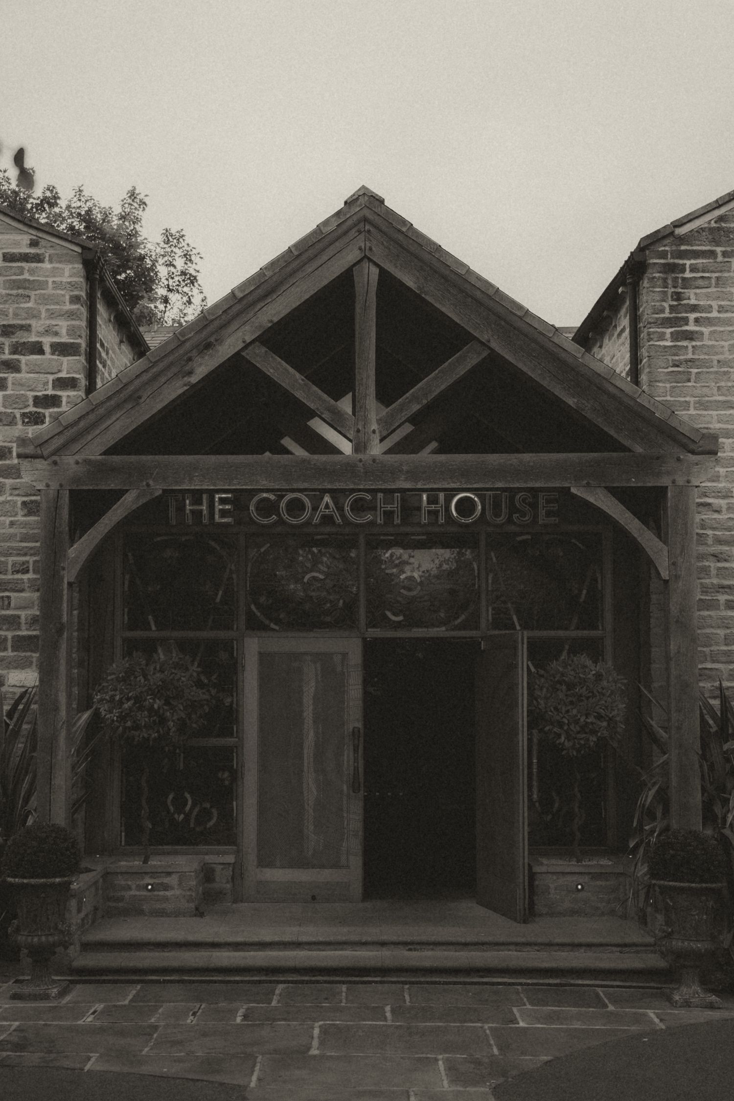 Black and white architectural photo of The Coach House entrance with brick walls and wooden beams.