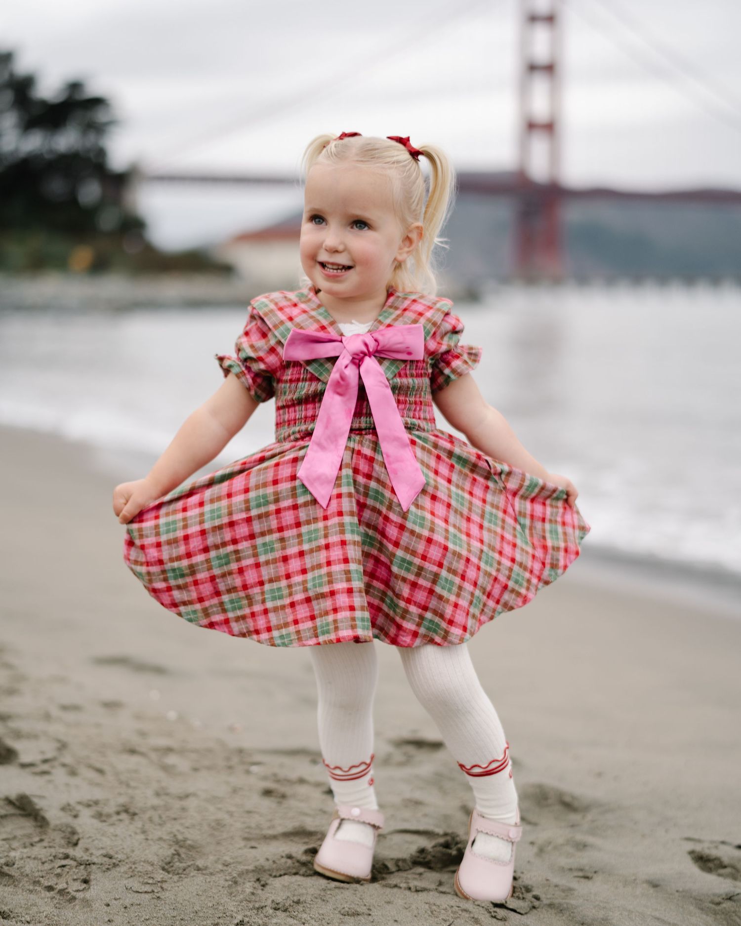 A young child in a plaid dress and white tights twirls on the beach with the Golden Gate Bridge in the background.