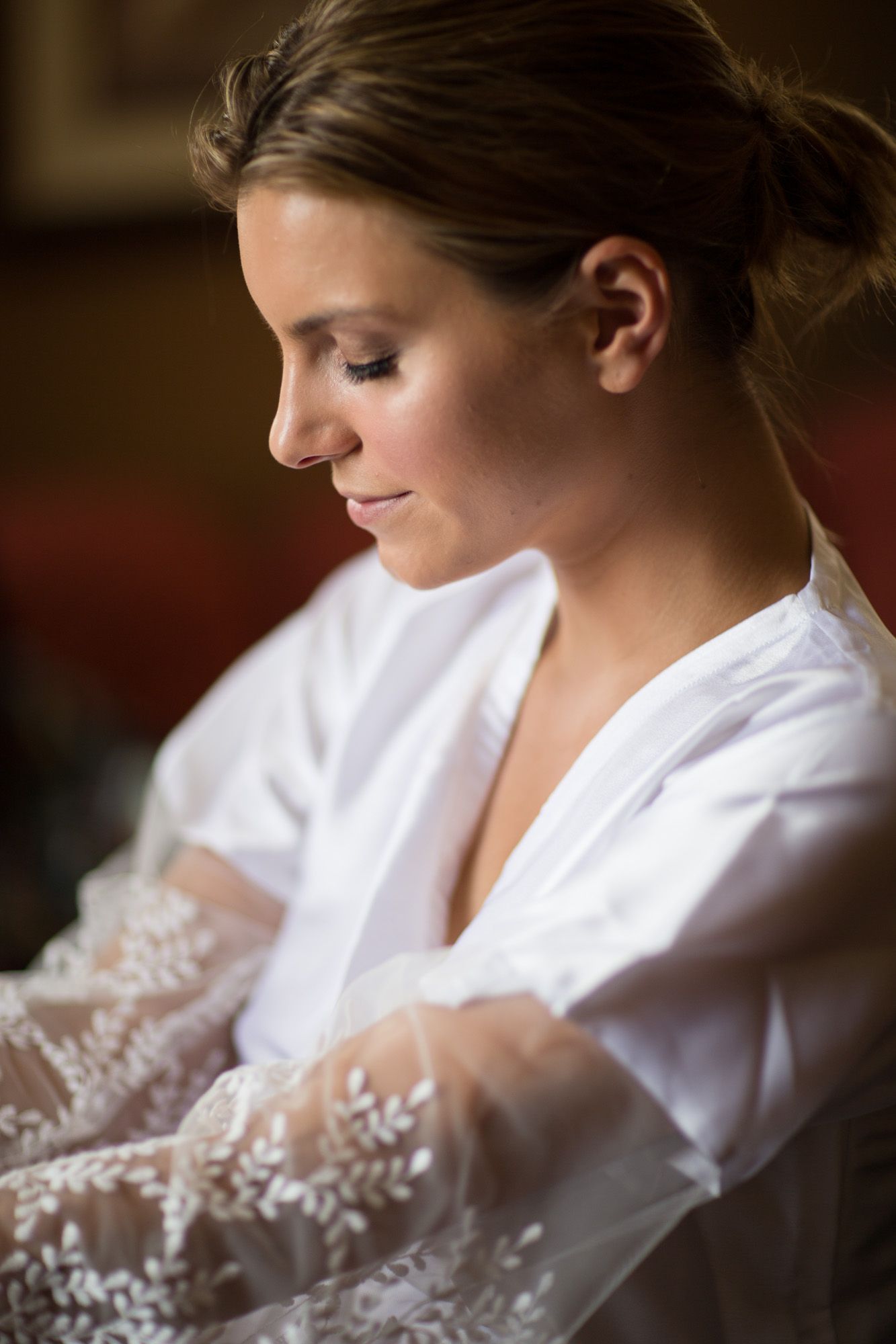 A bride in a white robe sits quietly while preparing for her wedding day, with delicate lace details visible.