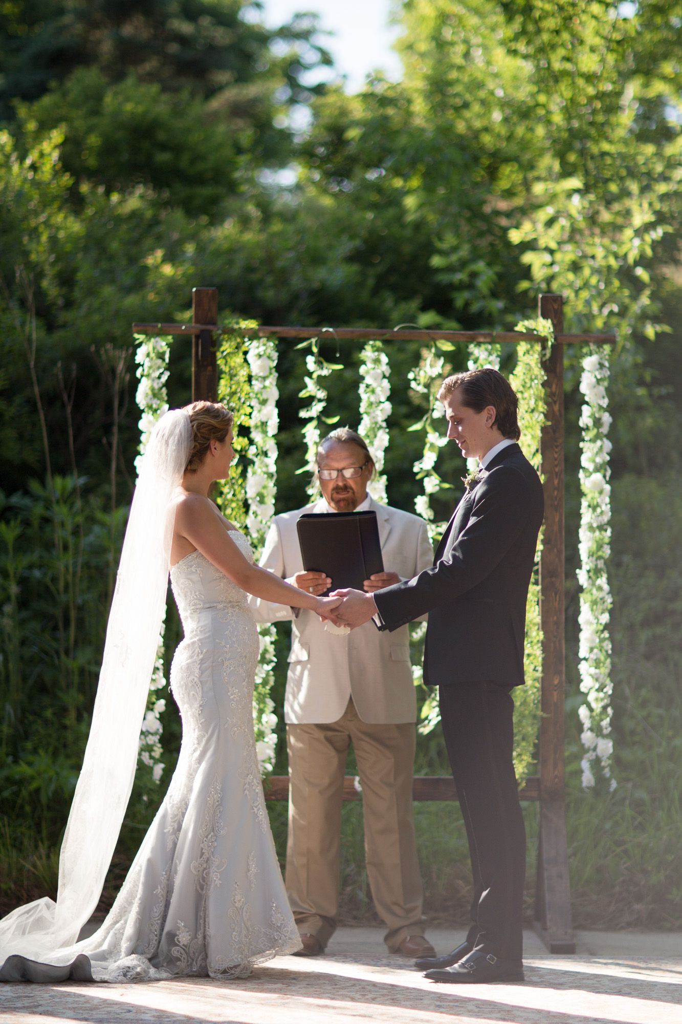 Wedding ceremony takes place outdoors under a white gazebo with greenery in the background.
