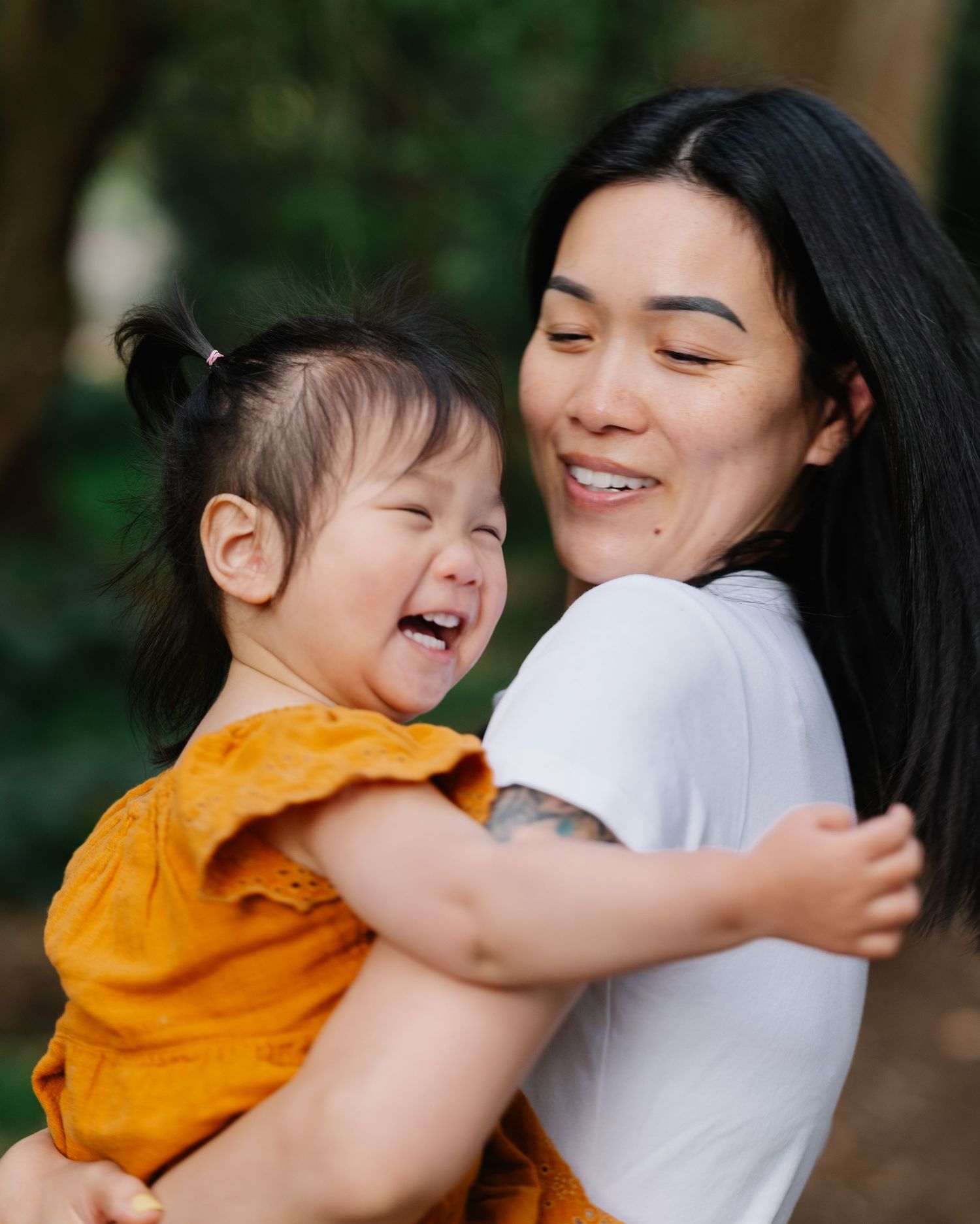 A smiling Asian woman holds a laughing toddler girl in an orange dress outdoors among green trees.