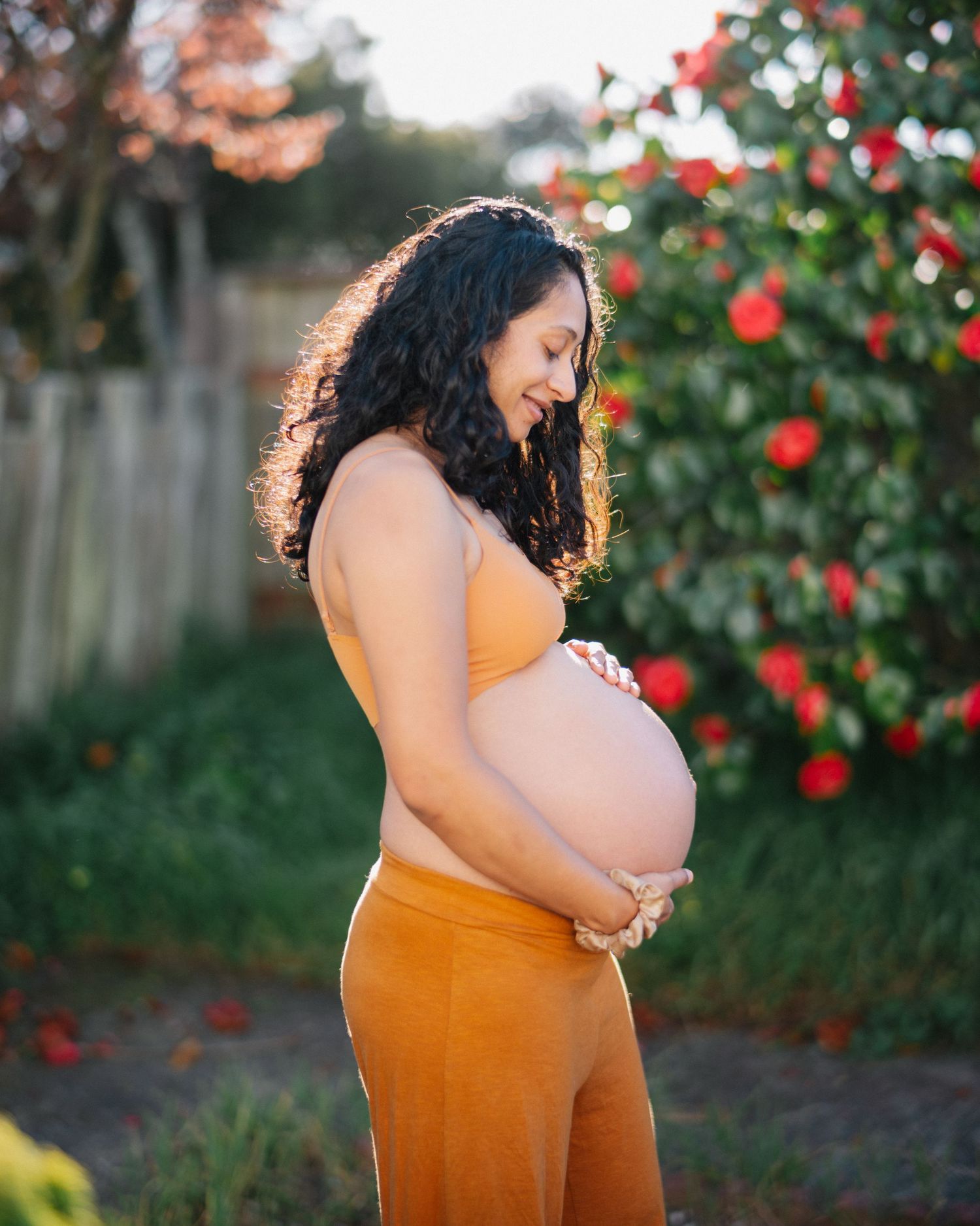 Pregnant woman in orange outfit cradling baby bump outdoors near red roses, smiling down at belly in golden light.